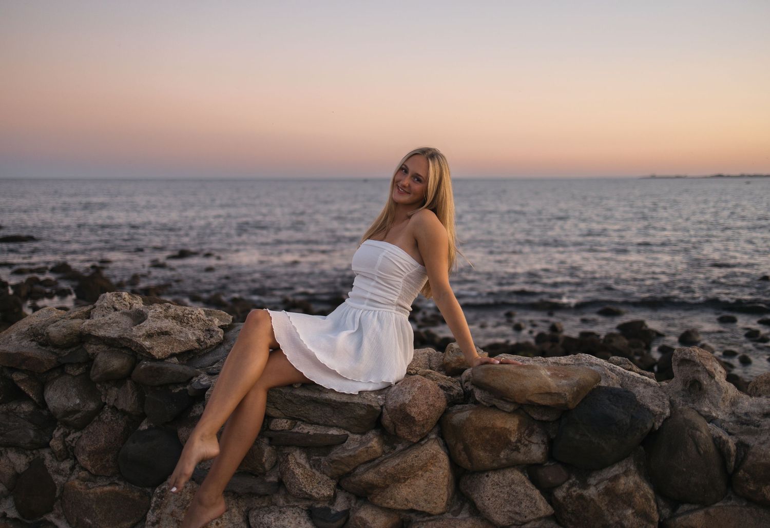 Someone in a white dress sits on coastal rocks during golden hour as waves crash in the background.