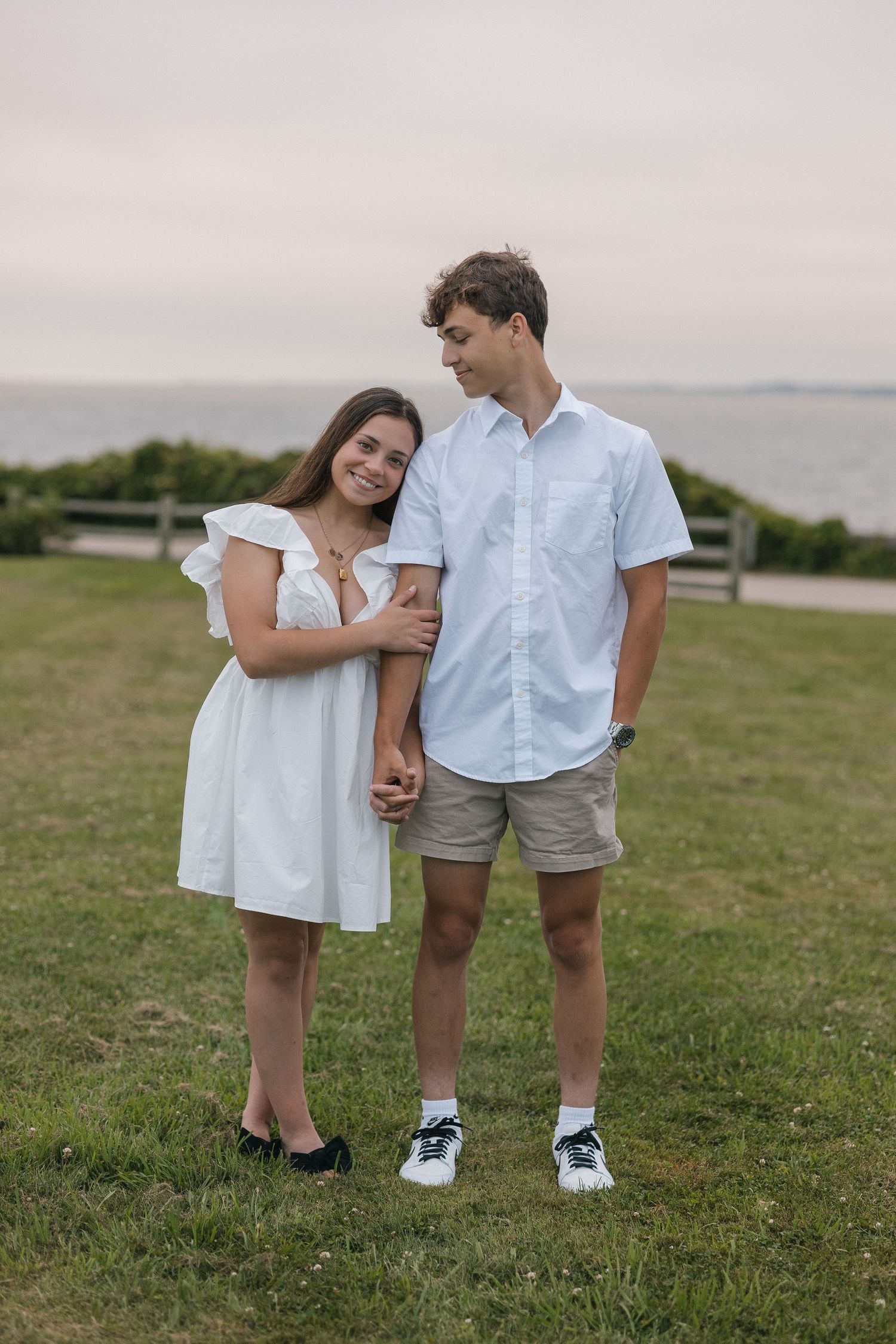 A couple in coordinated white outfits share a tender moment together on a grassy hilltop overlooking the ocean.