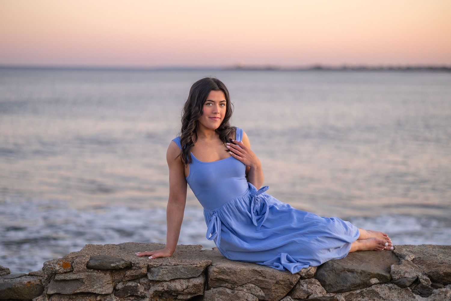 Woman in blue dress sitting on stone wall by the ocean at sunset.