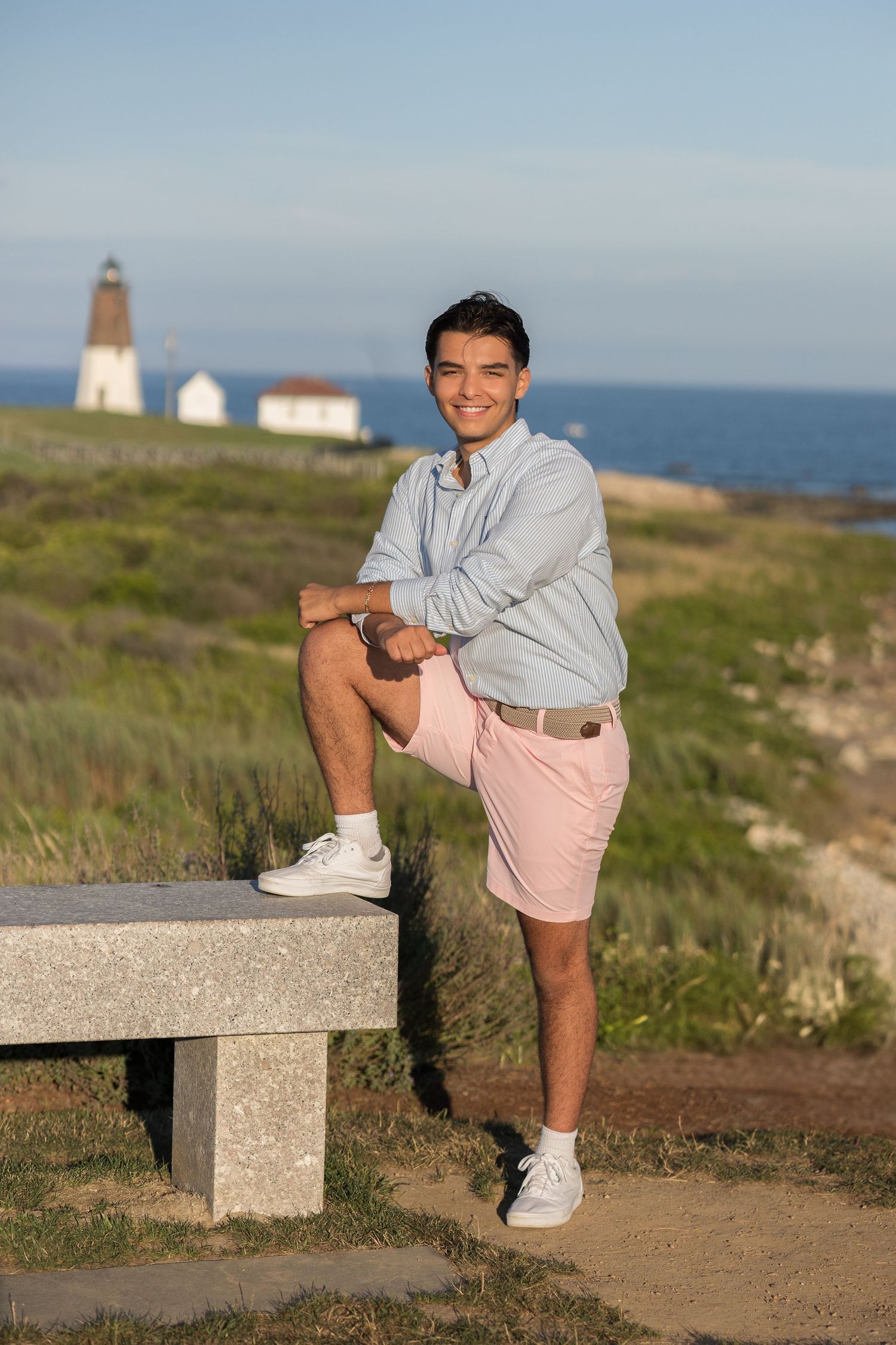 Person in casual summer attire poses near a lighthouse on coastal cliffside during golden hour.