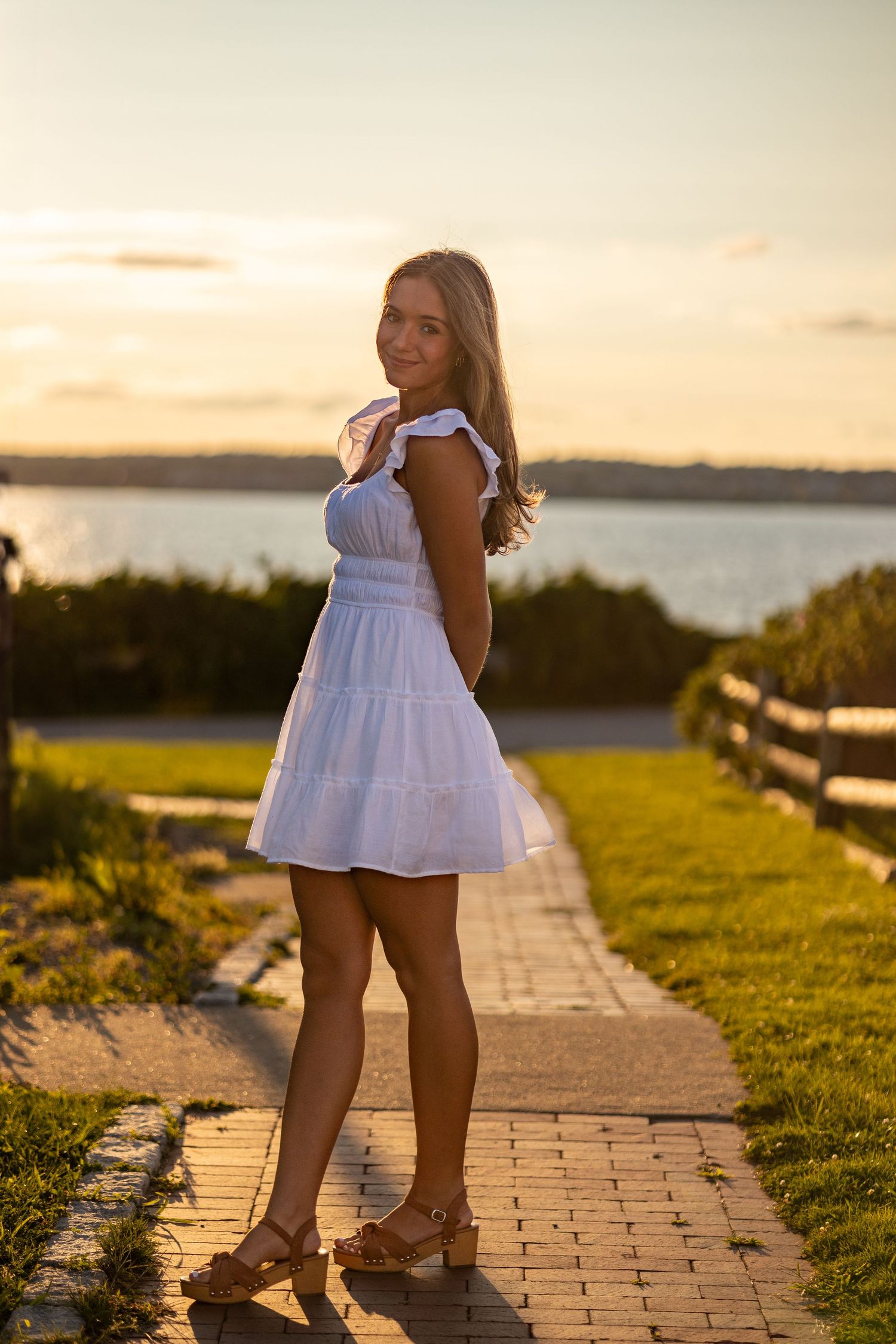 Woman in white summer dress poses on brick path during sunset by coastal overlook.