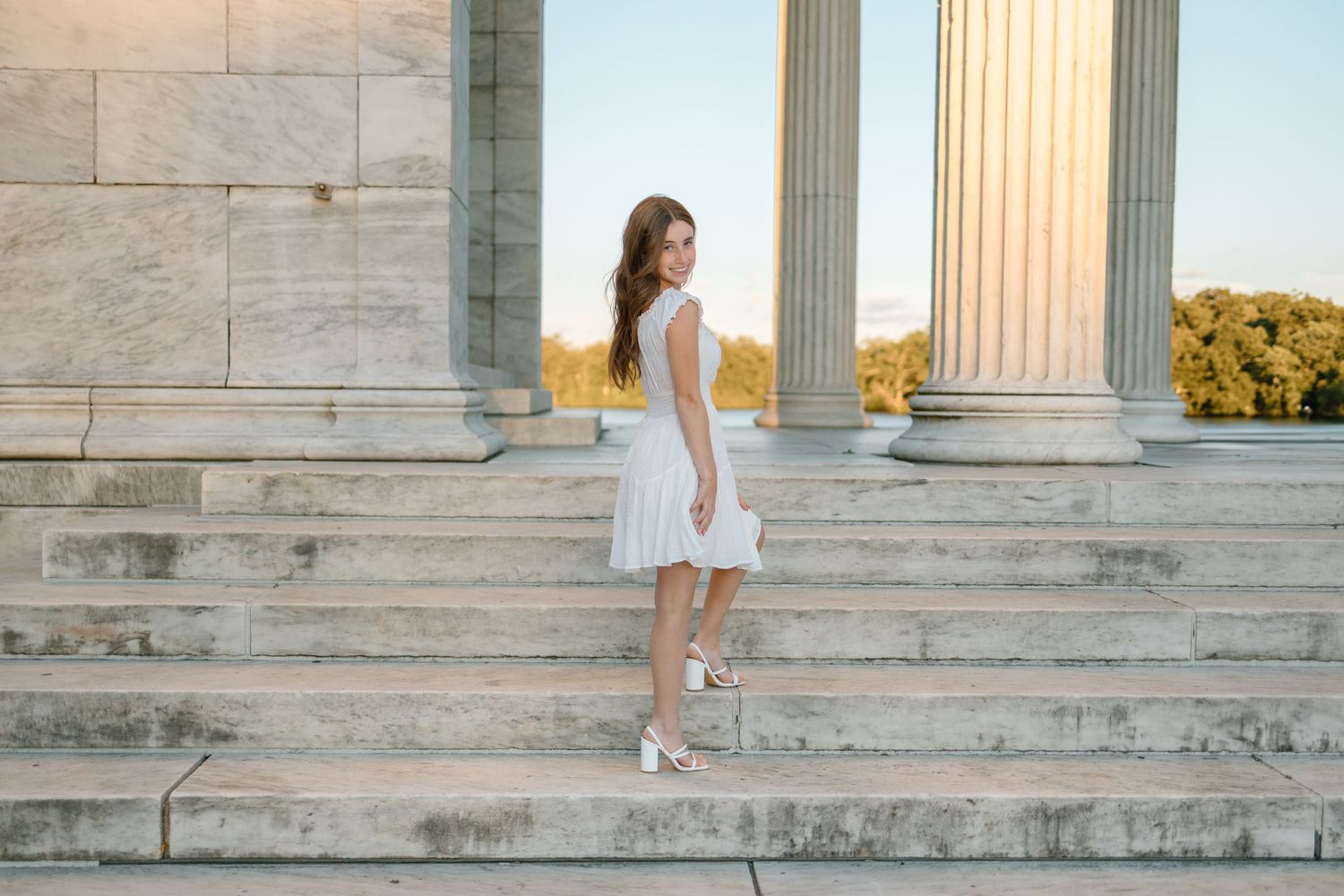 Woman in white dress stands on stone steps of classical building during sunset photoshoot.