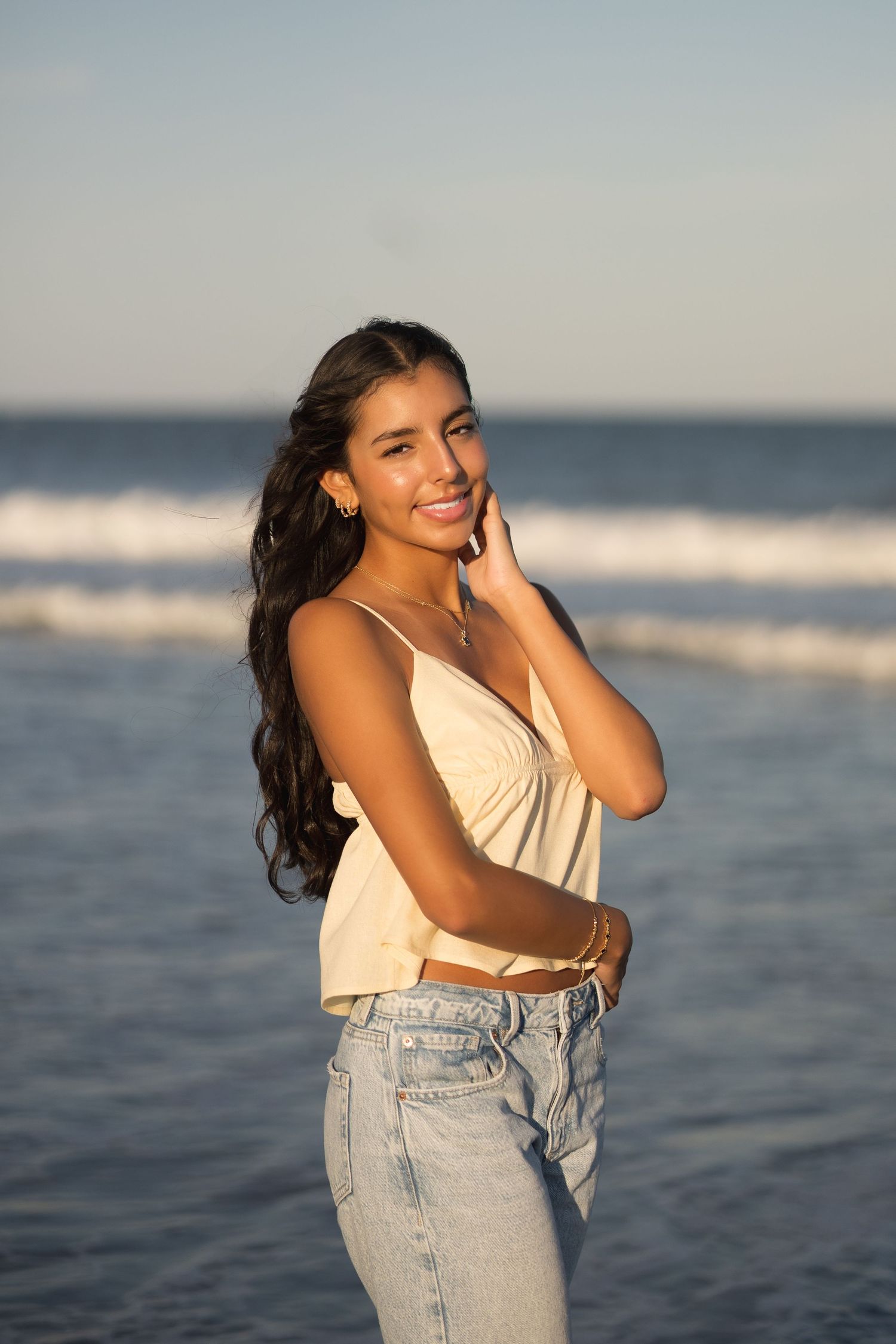 A person poses at the beach in Narragansett RI during golden hour wearing a cream top and light wash jeans with ocean waves in background.