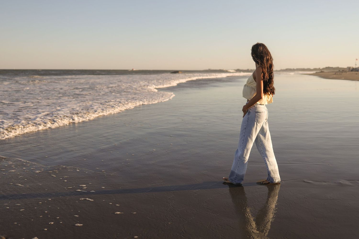 Person in white dress walks along calm beach shoreline at sunset creating reflections in wet sand.