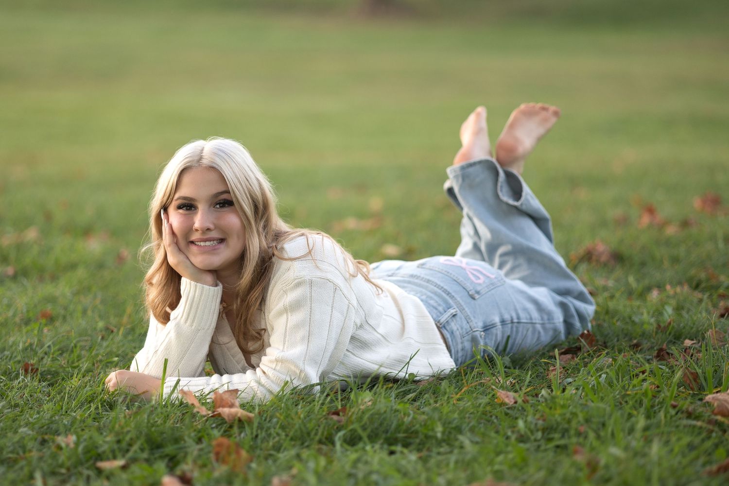 A person in white clothing lies on green grass during a sunny autumn day with scattered leaves around them.