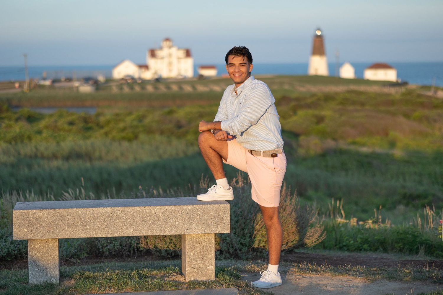 Someone poses on a stone bench near a lighthouse and coastal buildings during golden hour.