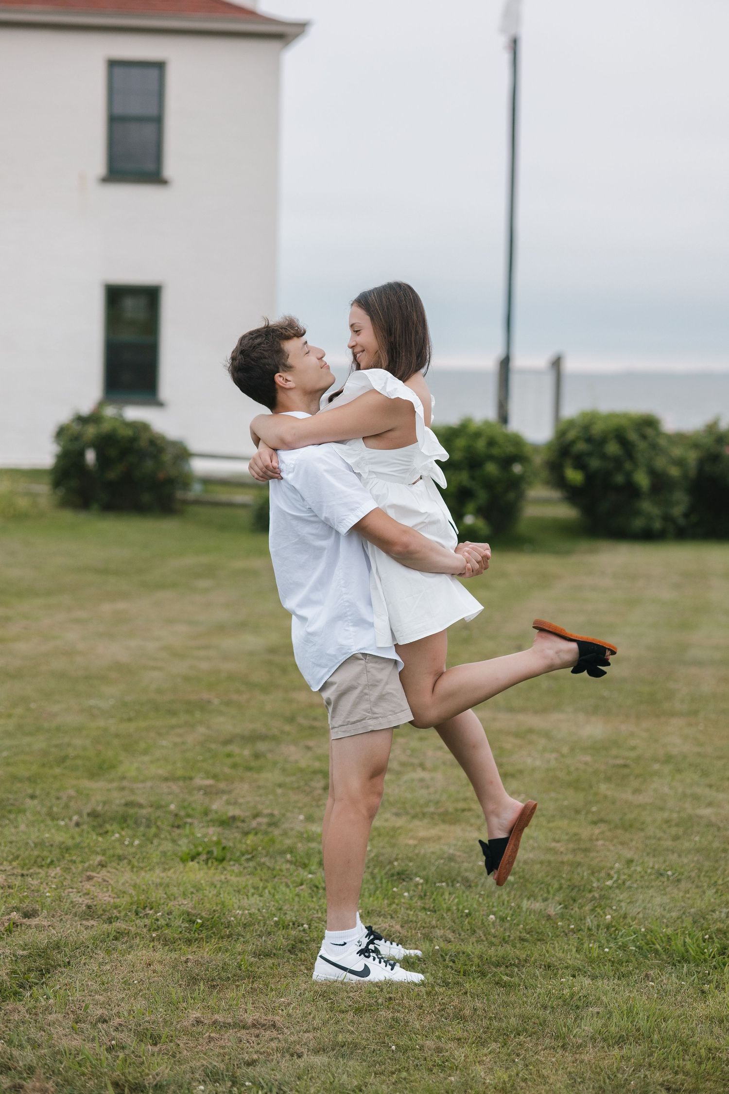 A couple dressed in white and khaki enjoys a lighthearted moment on a lawn near a white building.