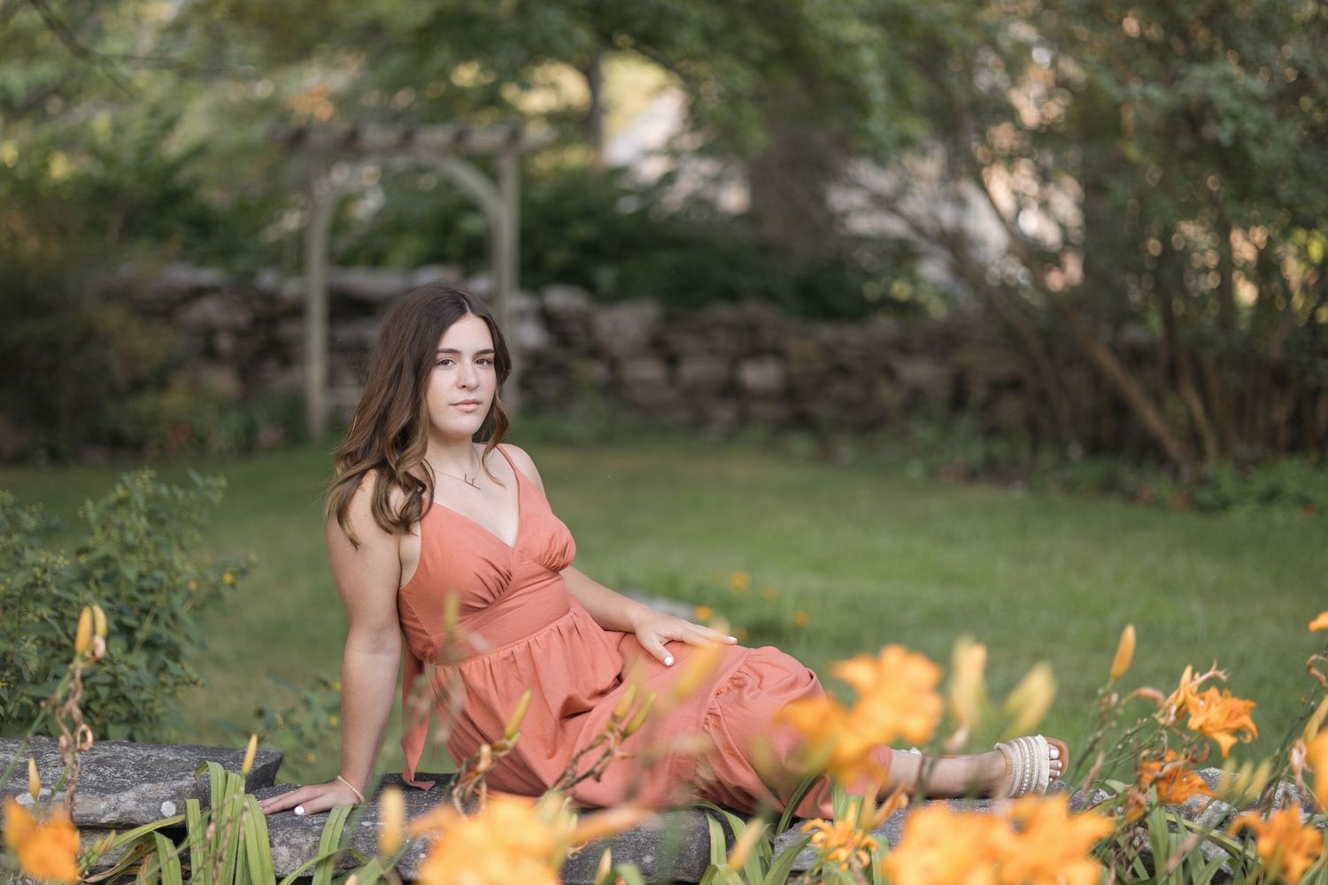 A woman in a peach-colored dress sits gracefully in a garden surrounded by blooming orange lilies.