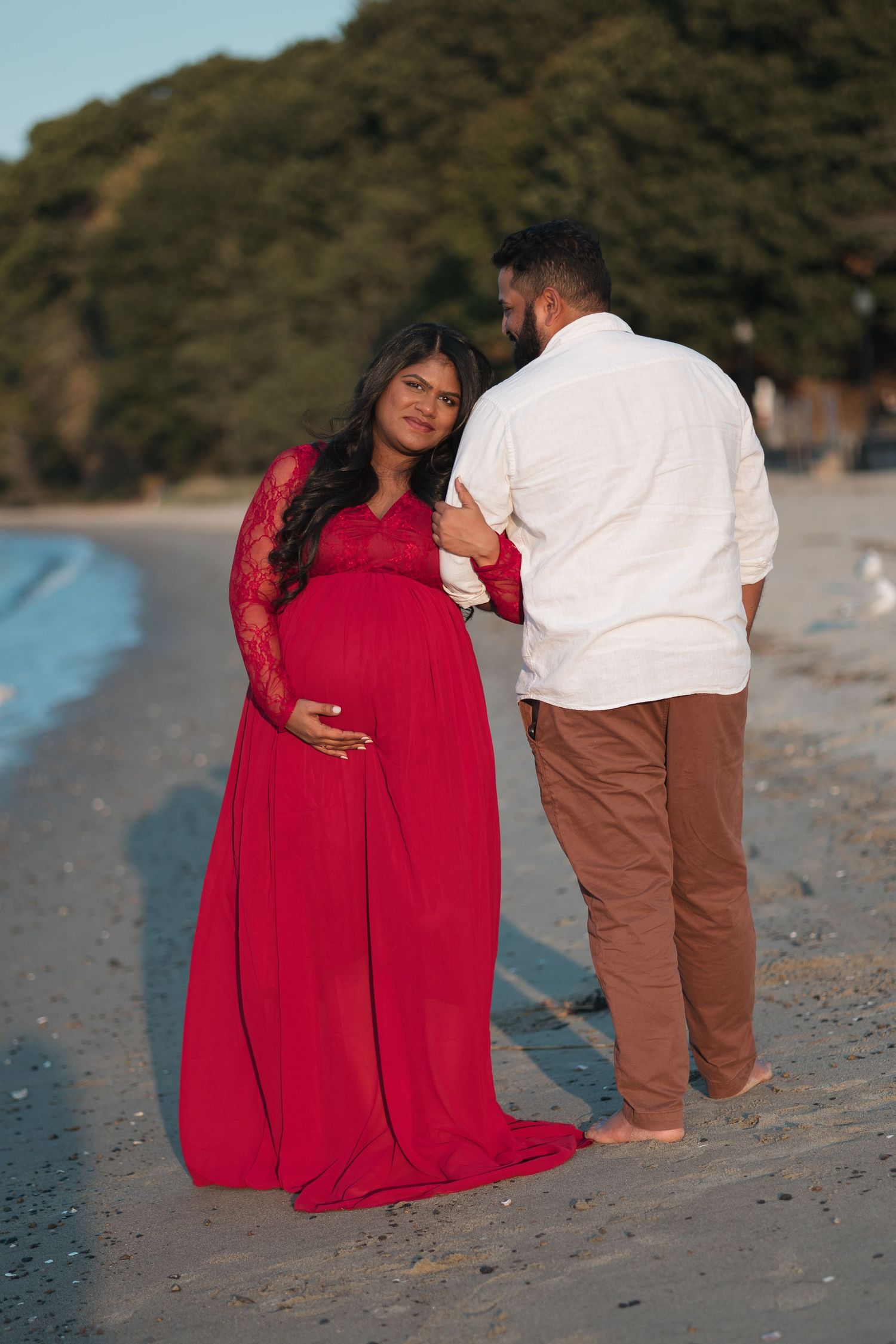 Expecting couple in red dress and white shirt pose on beach at sunset.
