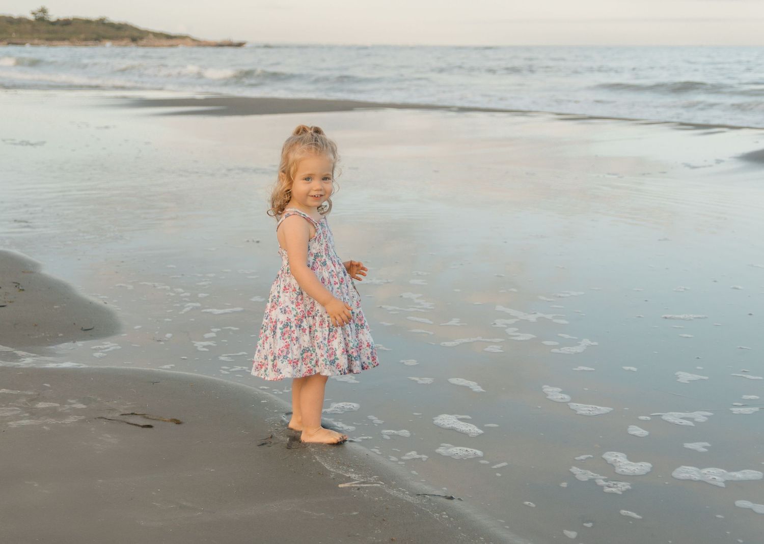 A toddler in a floral dress explores the beach at sunset.