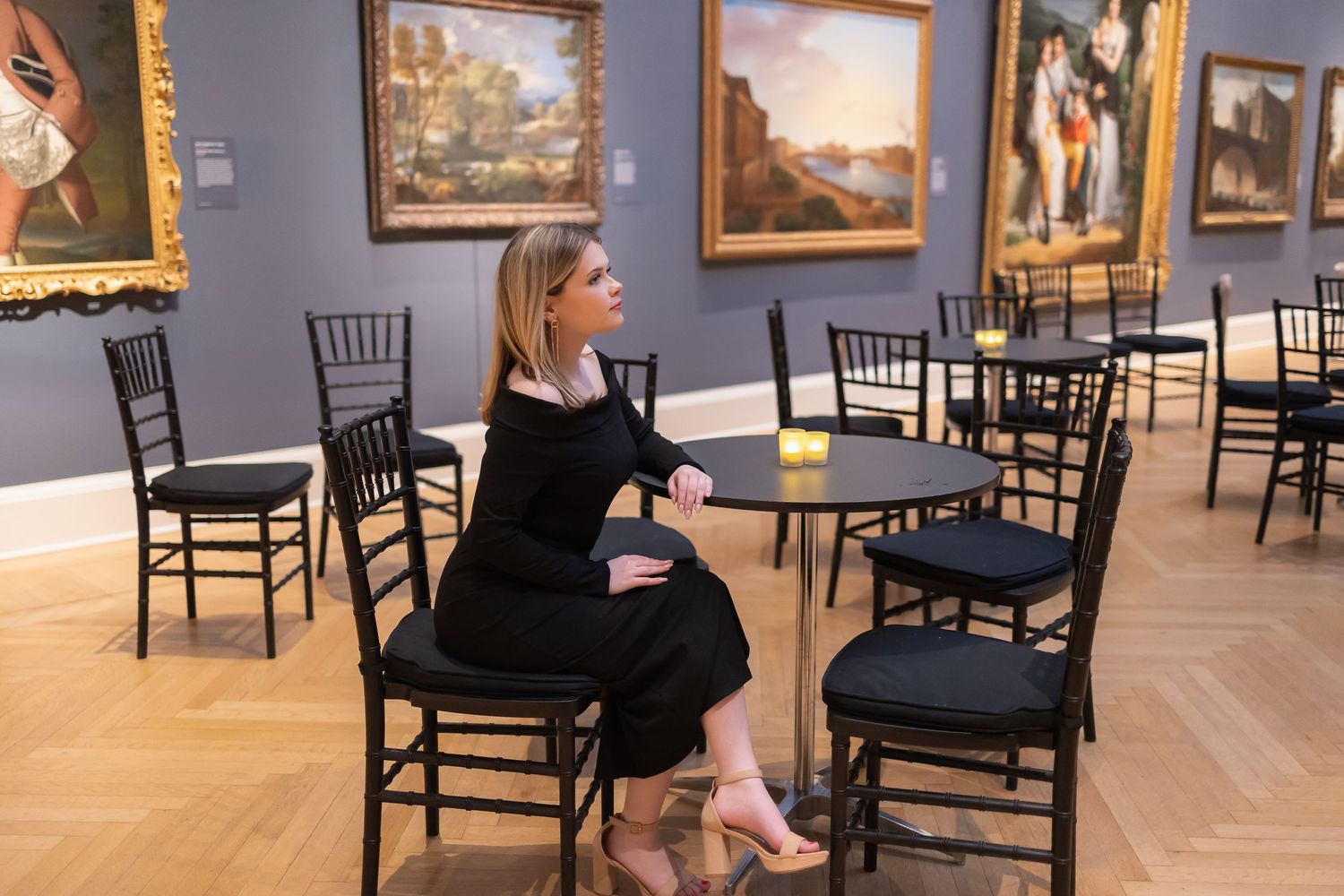 Person in black dress sits among empty chairs in museum gallery space.