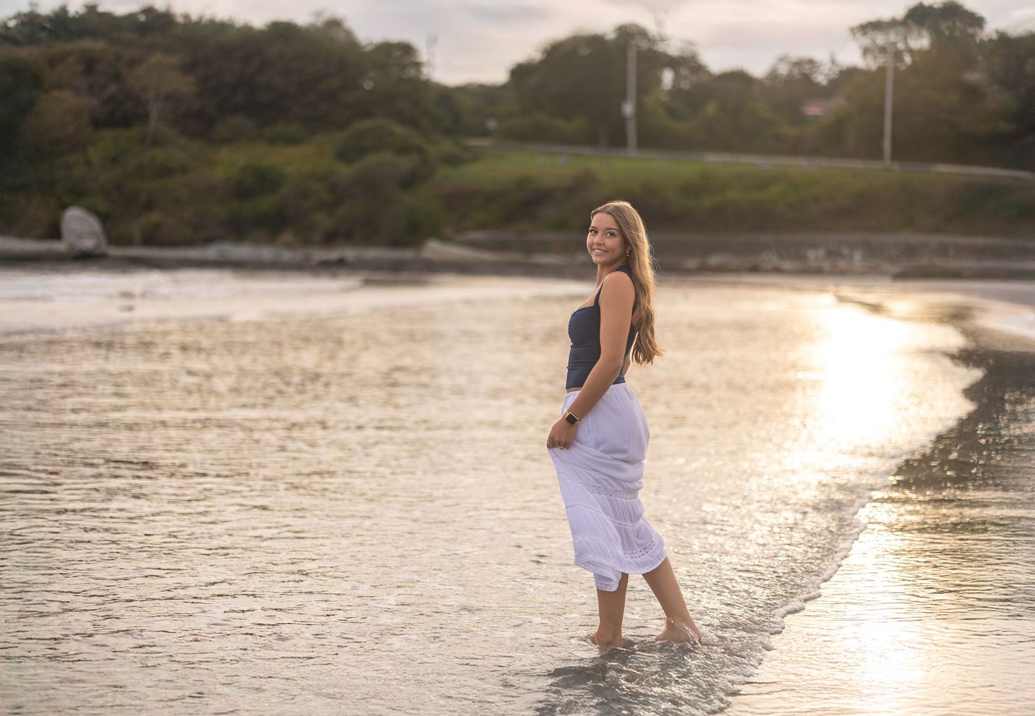 A High school senior walks through shallow ocean water in Newport Rhode Island at sunset creating rippling reflections on the calm surface.