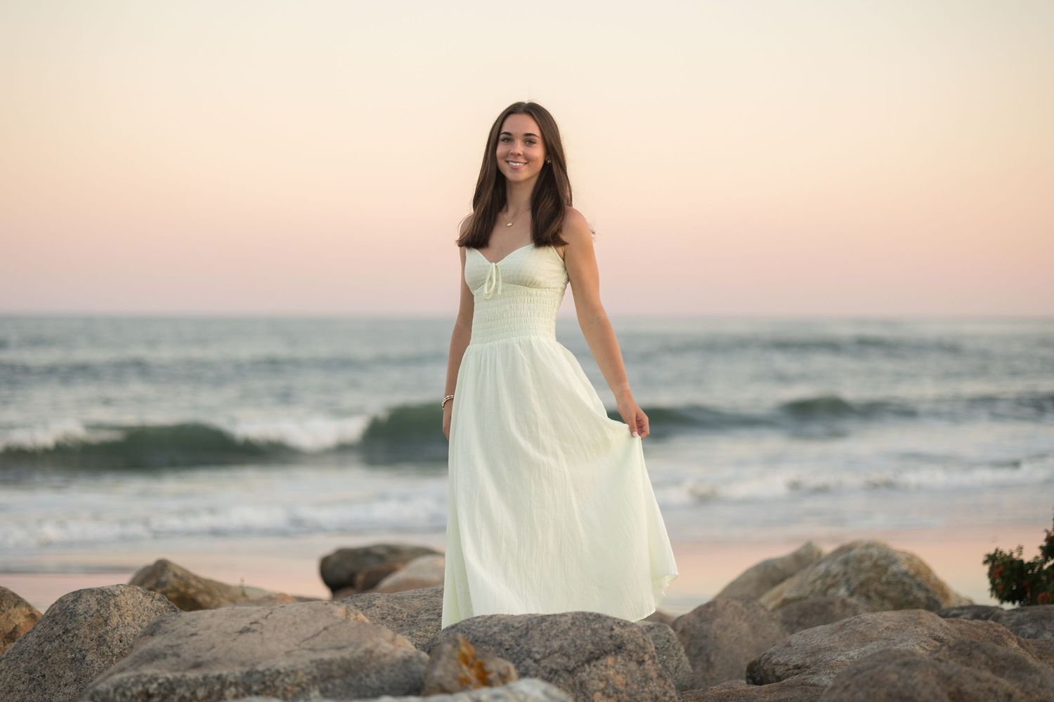 Woman in flowing white dress walks along rocky beach at sunset while ocean waves crash behind her.