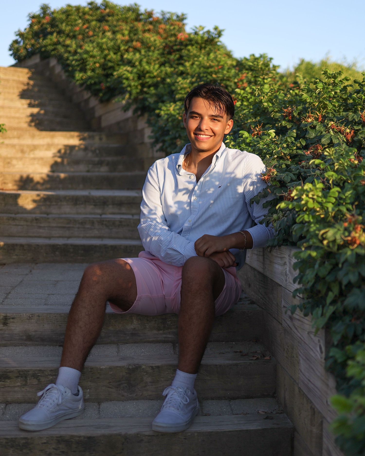 Person in white shirt and shorts sitting casually on stone steps with greenery in background during sunset.