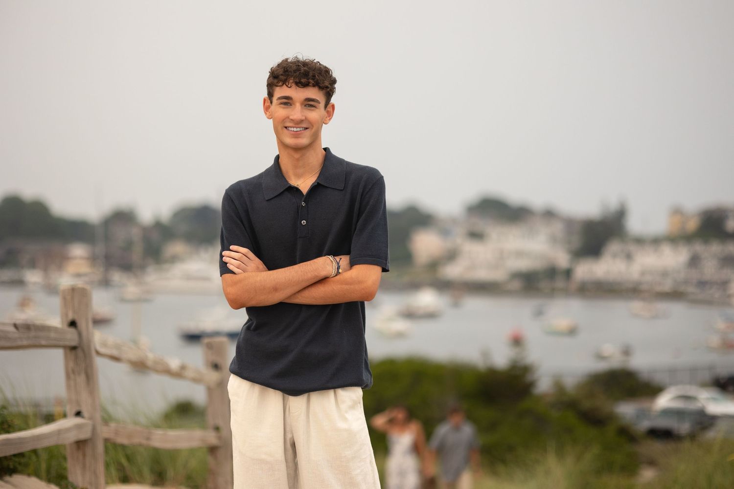 A person in a black shirt and khaki pants stands confidently by a coastal overlook with boats in the background.
