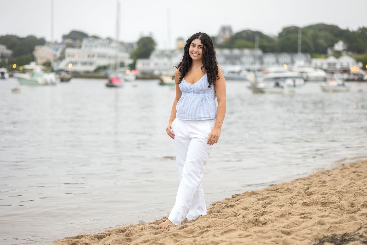 A woman in white clothing poses gracefully along a sandy beach with boats and water in the background.