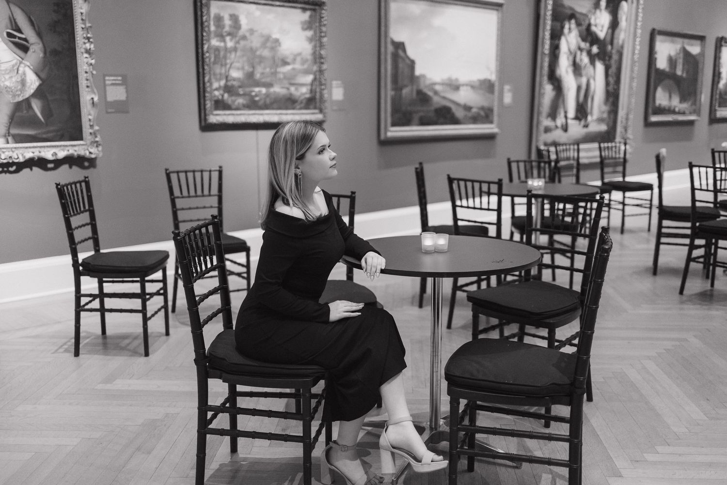 Black and white photo in an art gallery with chairs and framed paintings on display.