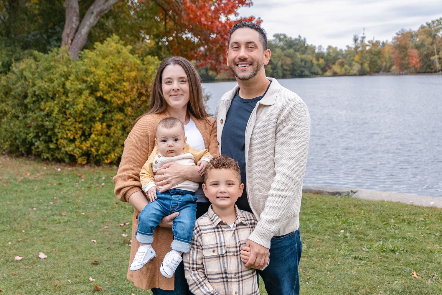 A family poses together on an autumn day by a lake with fall foliage in the background.