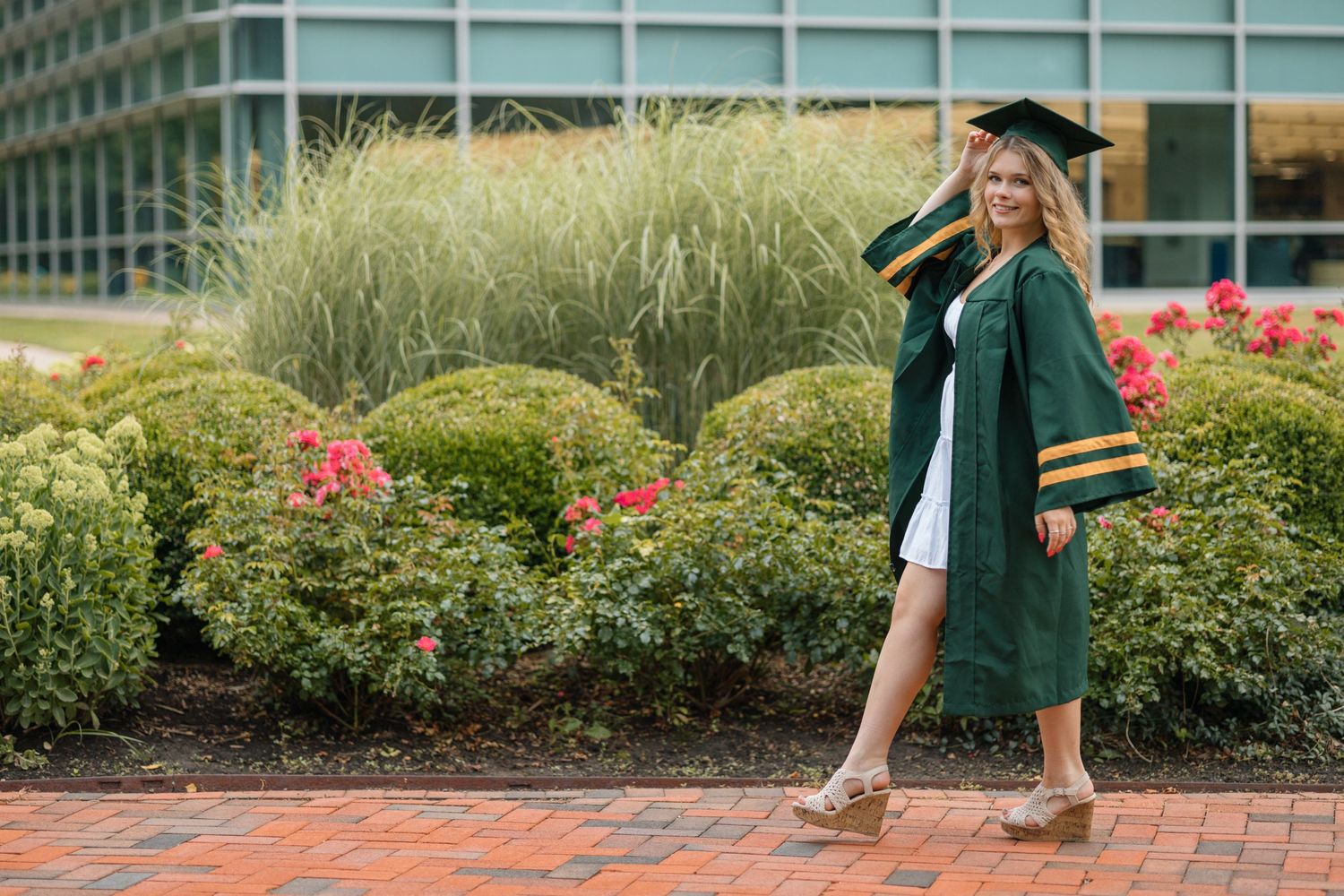 A graduate in green academic regalia strikes a pose on a brick pathway surrounded by landscaped gardens.