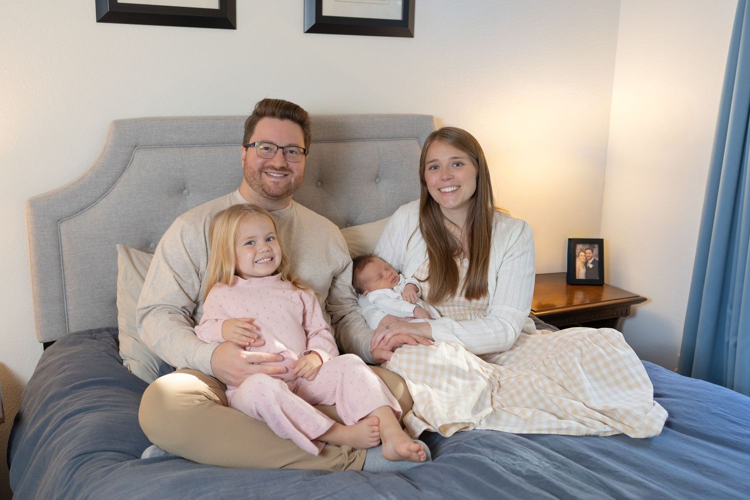 A family sits together on a gray upholstered bed, with two young children in casual home attire.