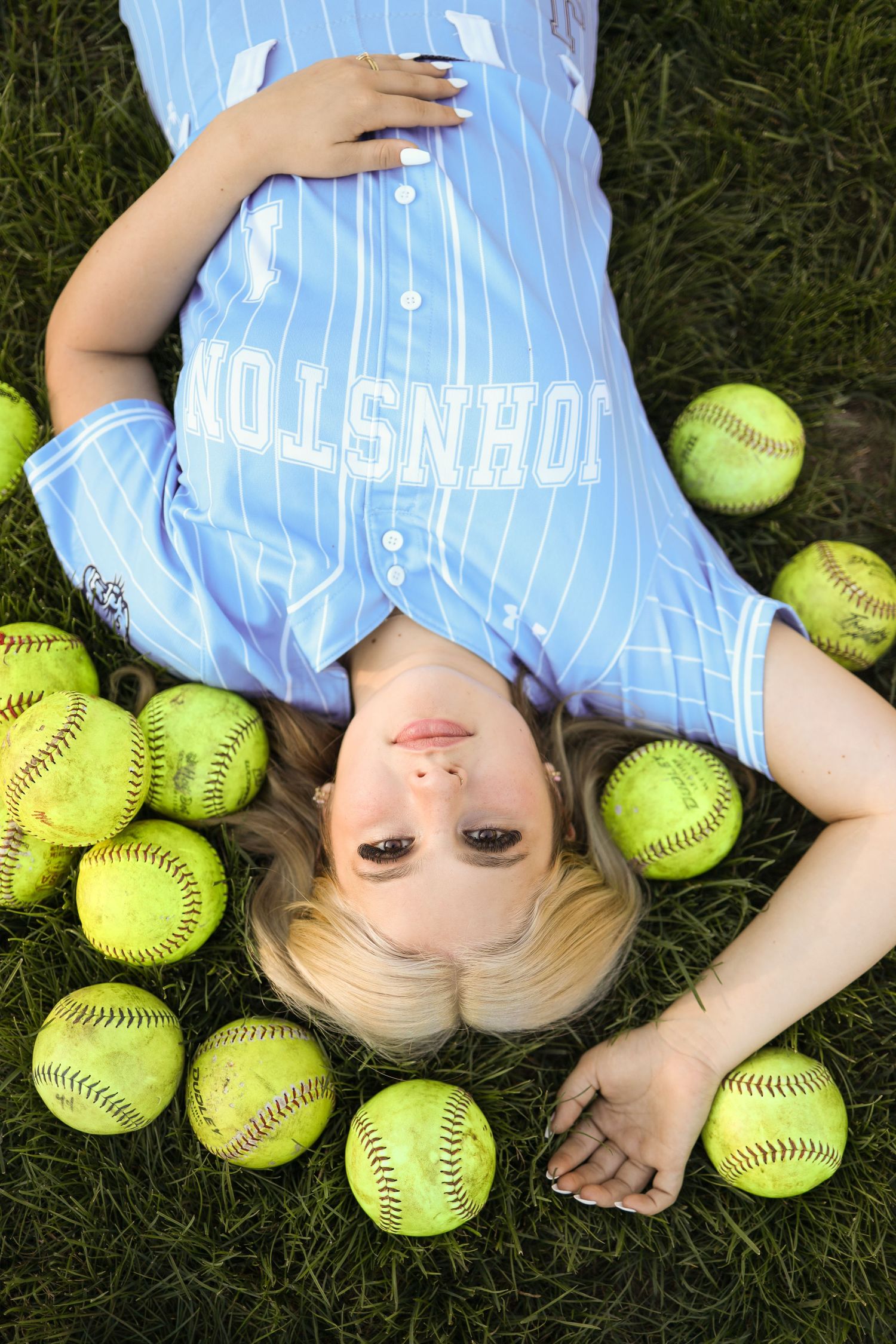 Softball player lying on grass surrounded by bright yellow softballs wearing a pinstripe uniform.