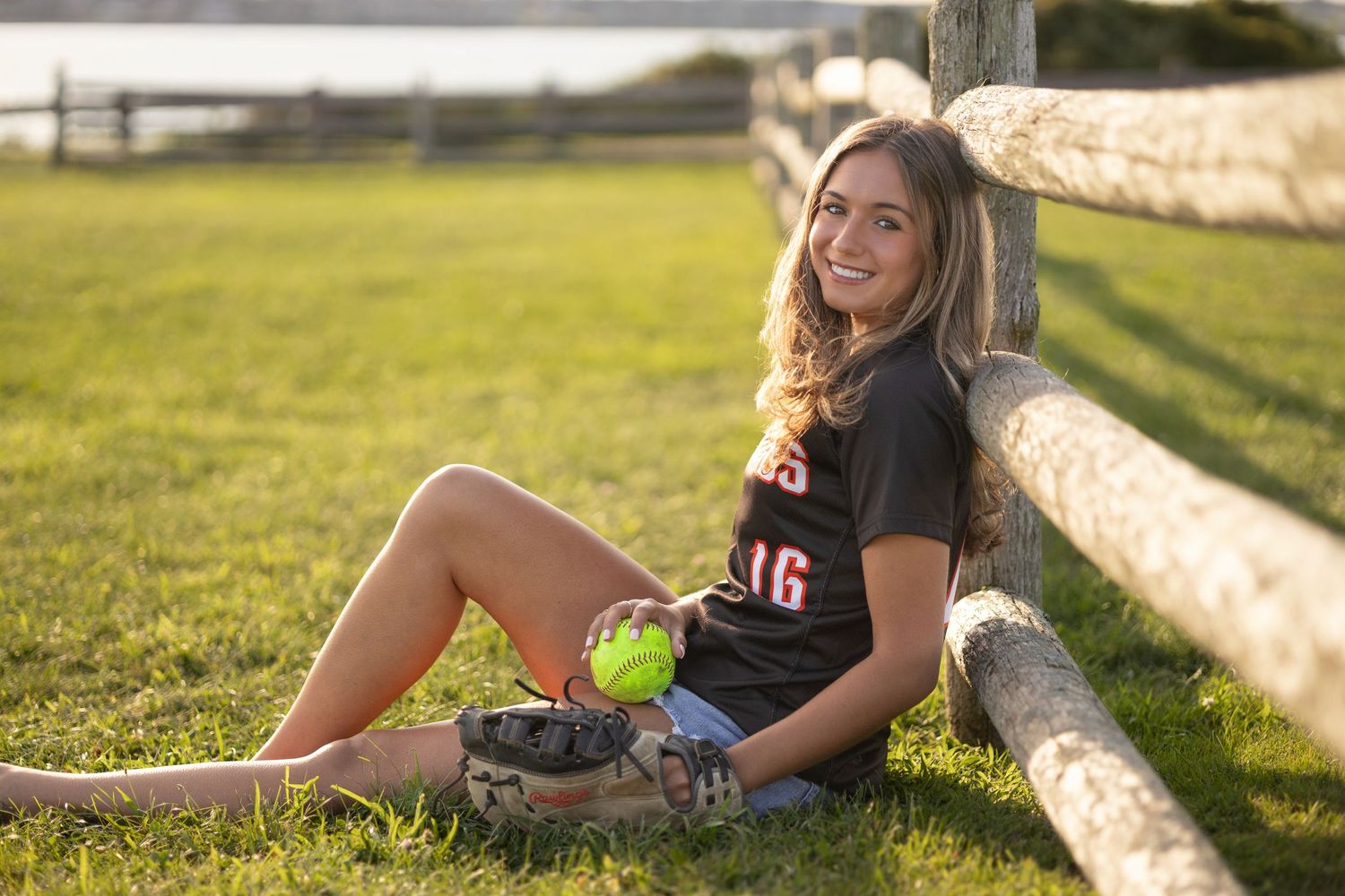 A tennis player relaxes on grass next to a white fence while holding a tennis ball.