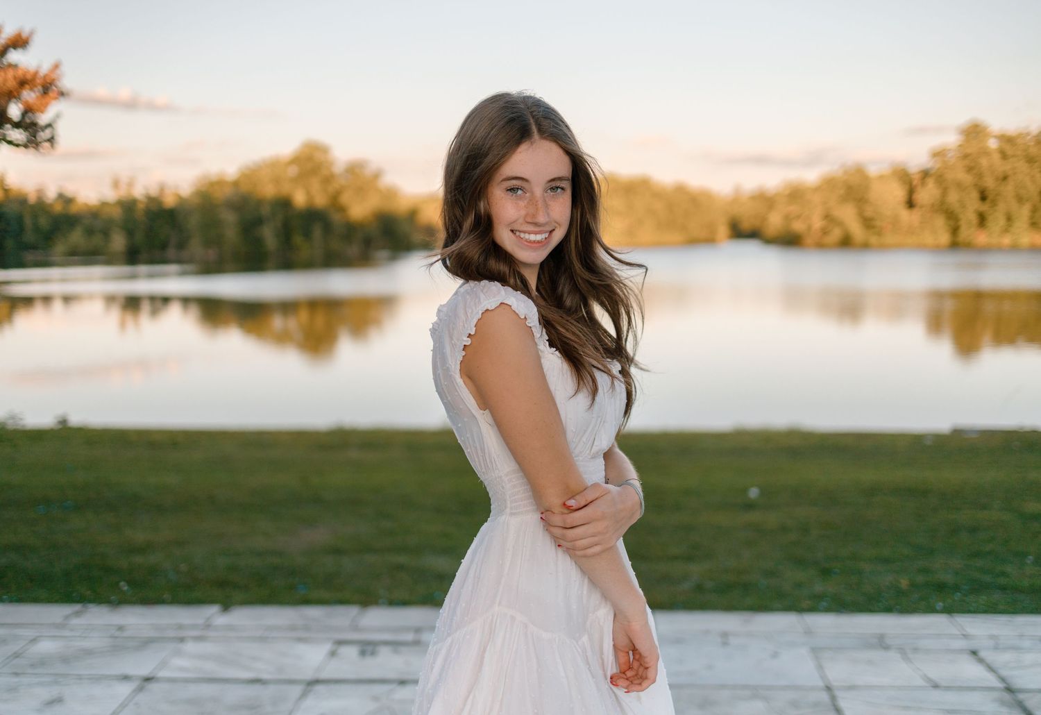 Woman in white dress poses gracefully on lakeside path during sunset portrait photography session.