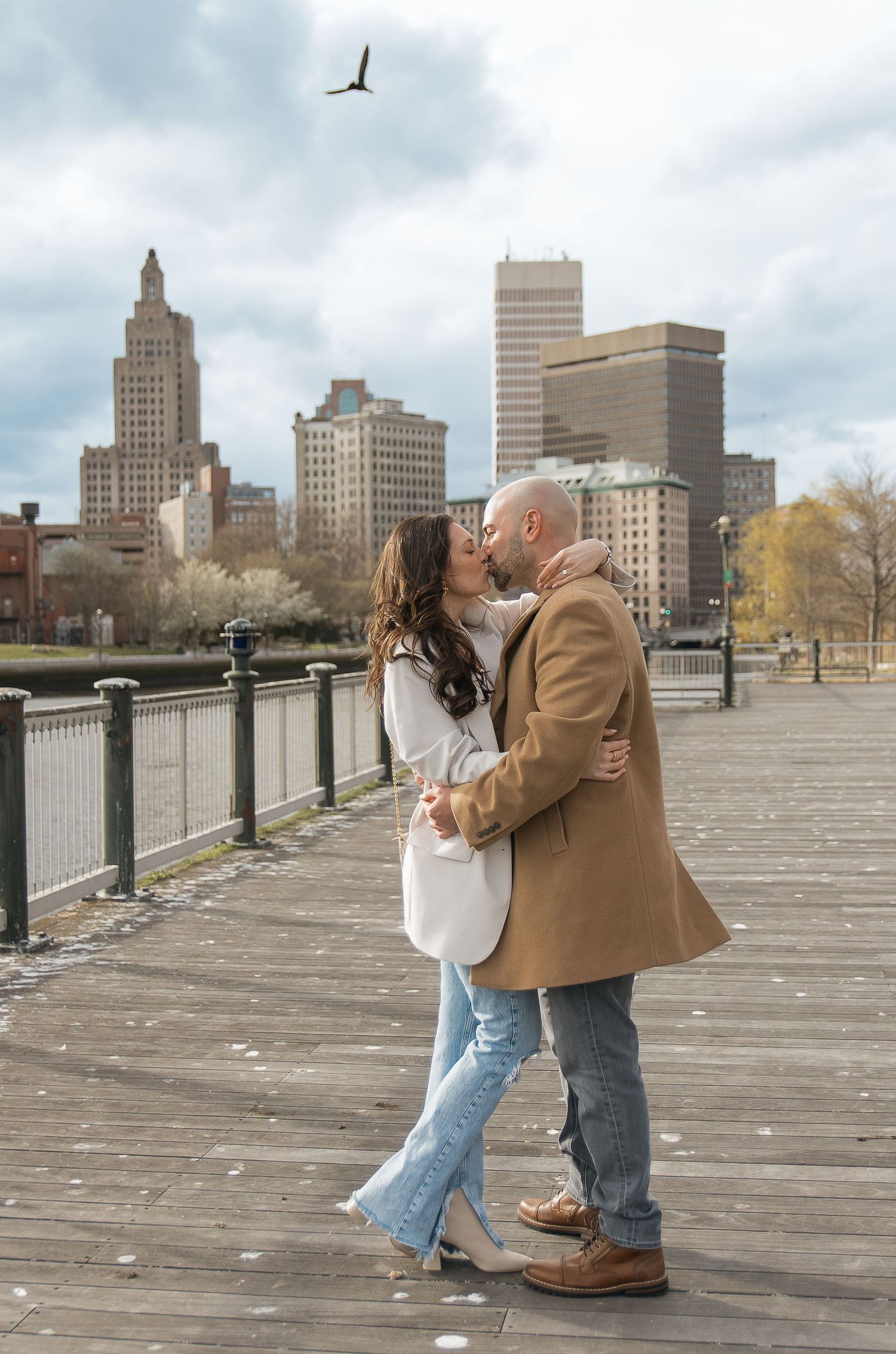A romantic moment on a boardwalk with Providence city skyline and a bird flying overhead.