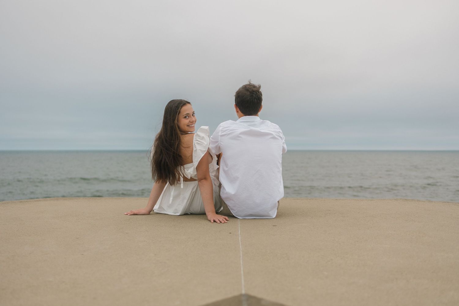 A couple sits together on a sandy beach overlooking the ocean on a cloudy day.