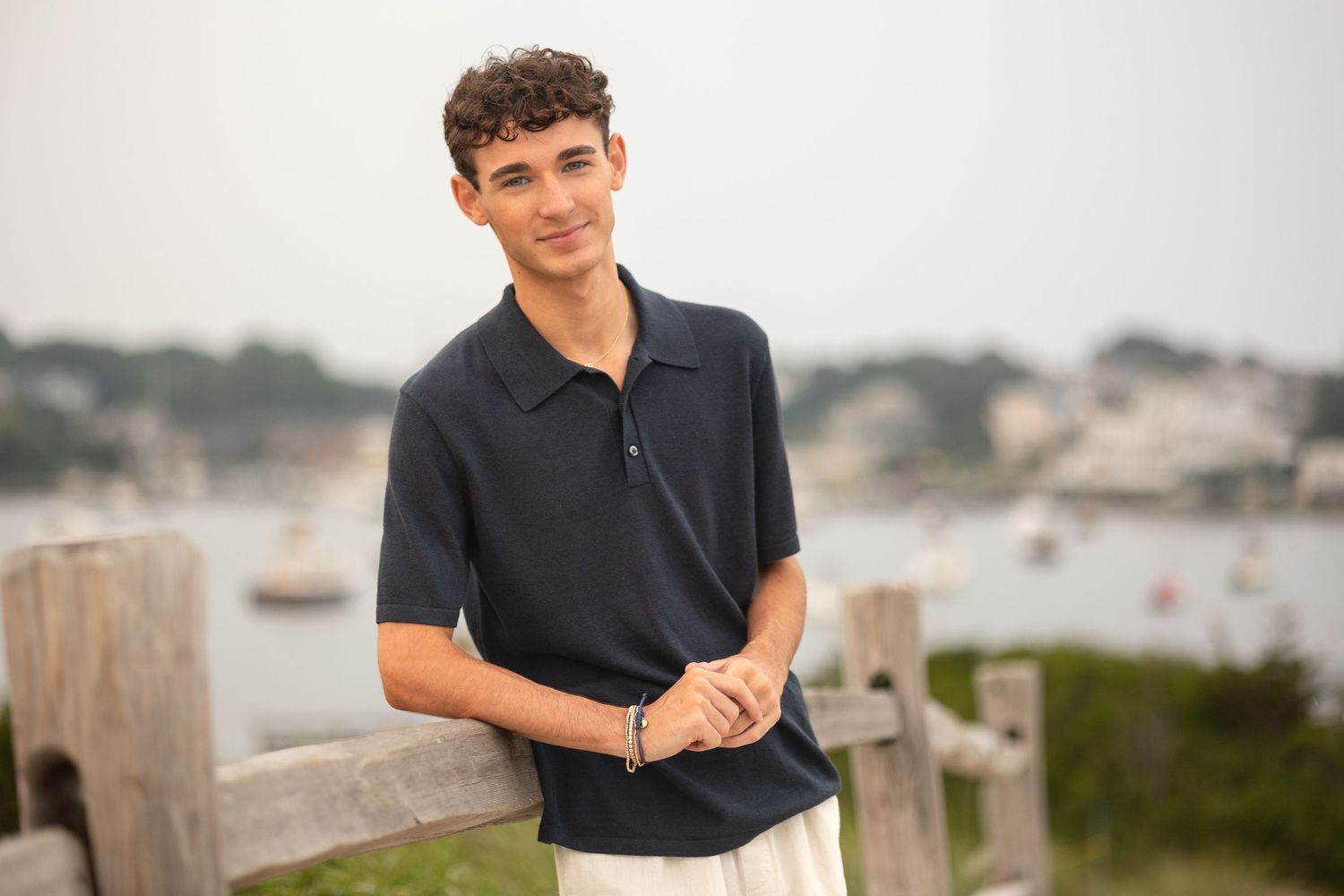 Young man in black polo shirt smiles while standing on wooden dock by waterfront at sunset.