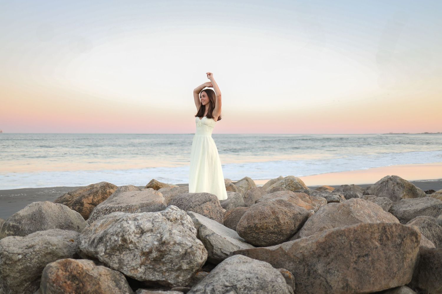 A person in a white dress stands on coastal rocks at sunset overlooking the ocean.