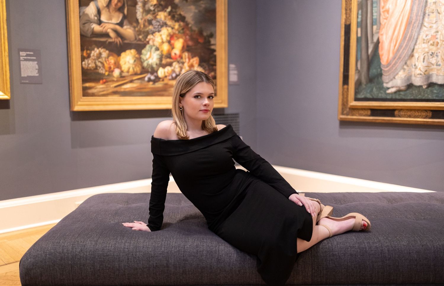 Figure in black dress poses on padded bench against gallery wall with artwork.