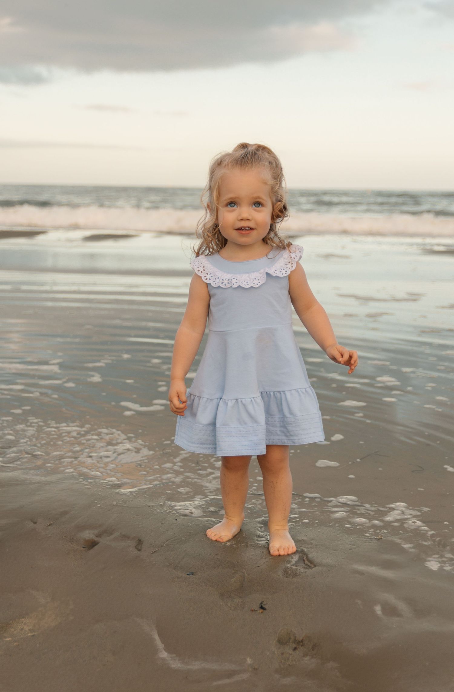 A toddler in a blue dress stands on the beach with waves and clouds in the background.