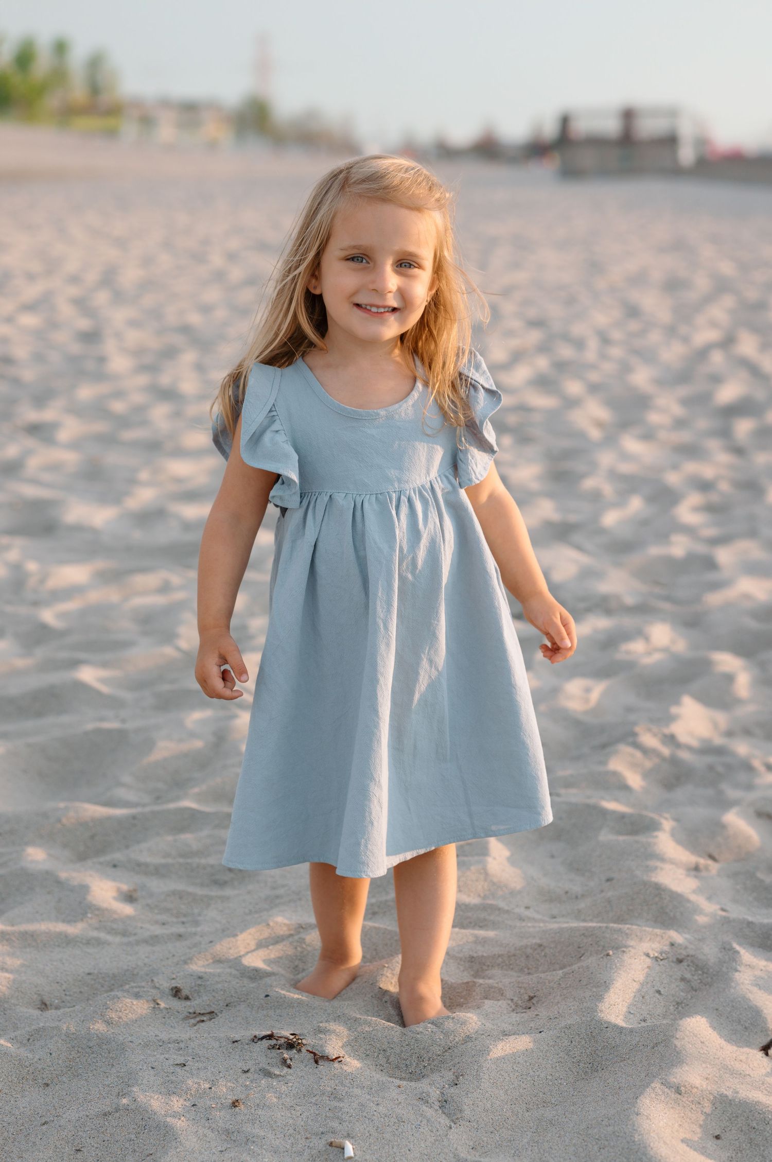 Young child in light blue dress standing barefoot on sandy beach.