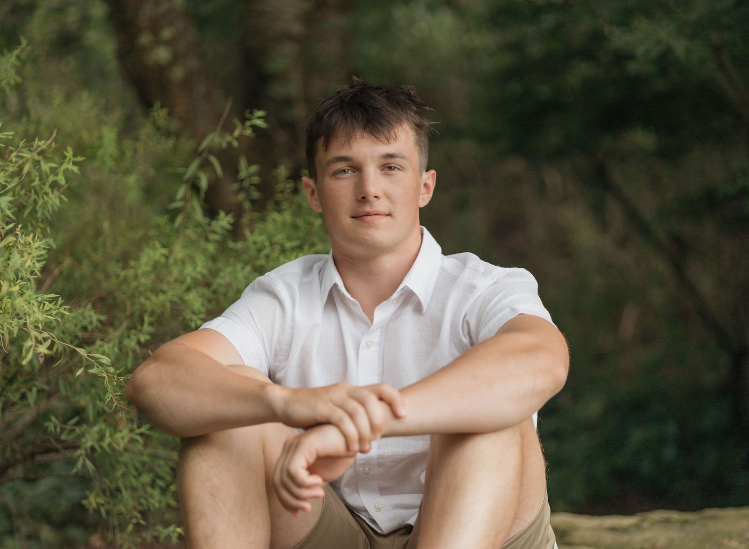 A person in a white shirt sits casually outdoors against a natural green background.