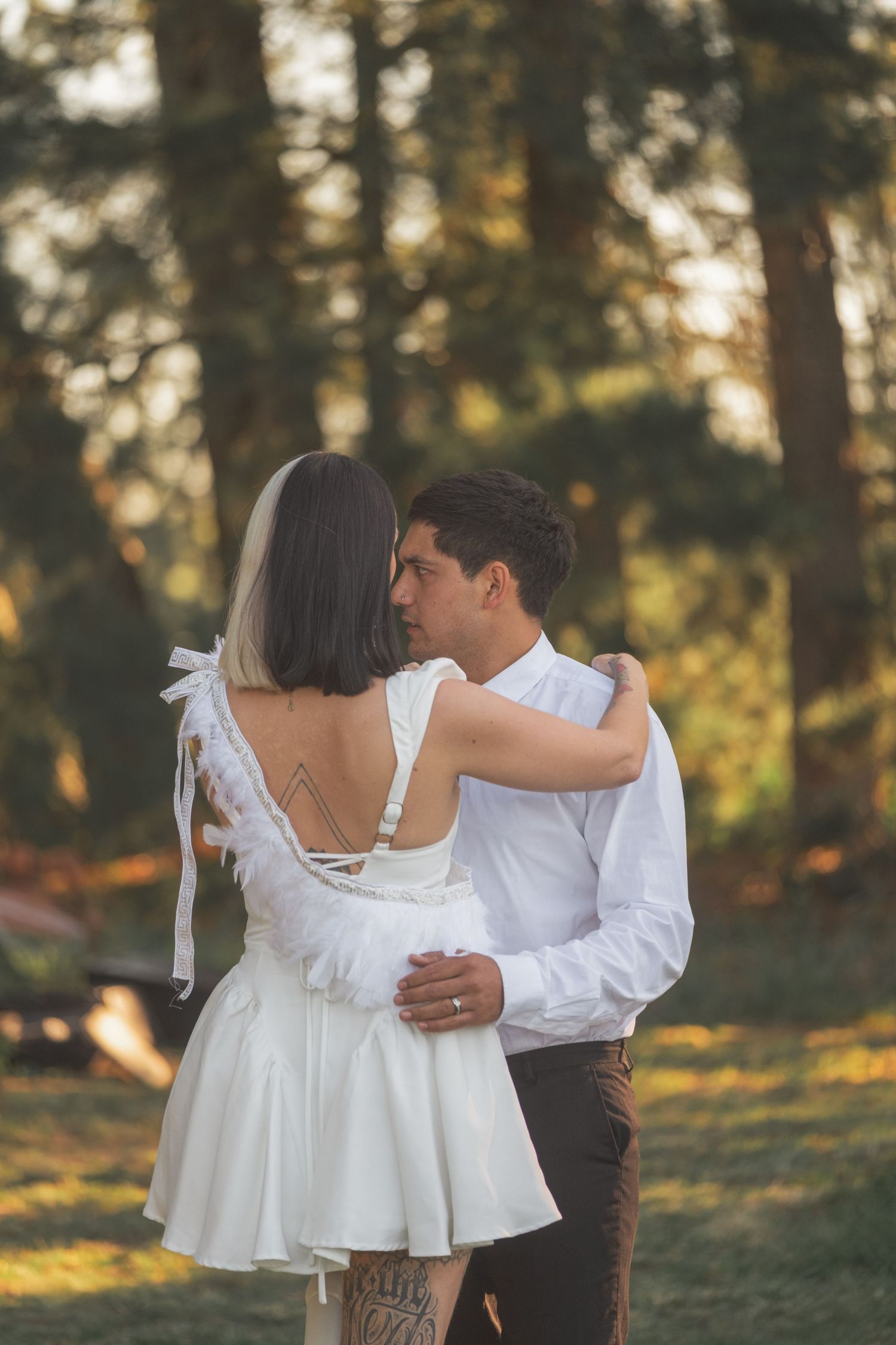 Couple embracing in a sunlit forest, bride in white backless mini dress with tattoo, groom in white shirt.