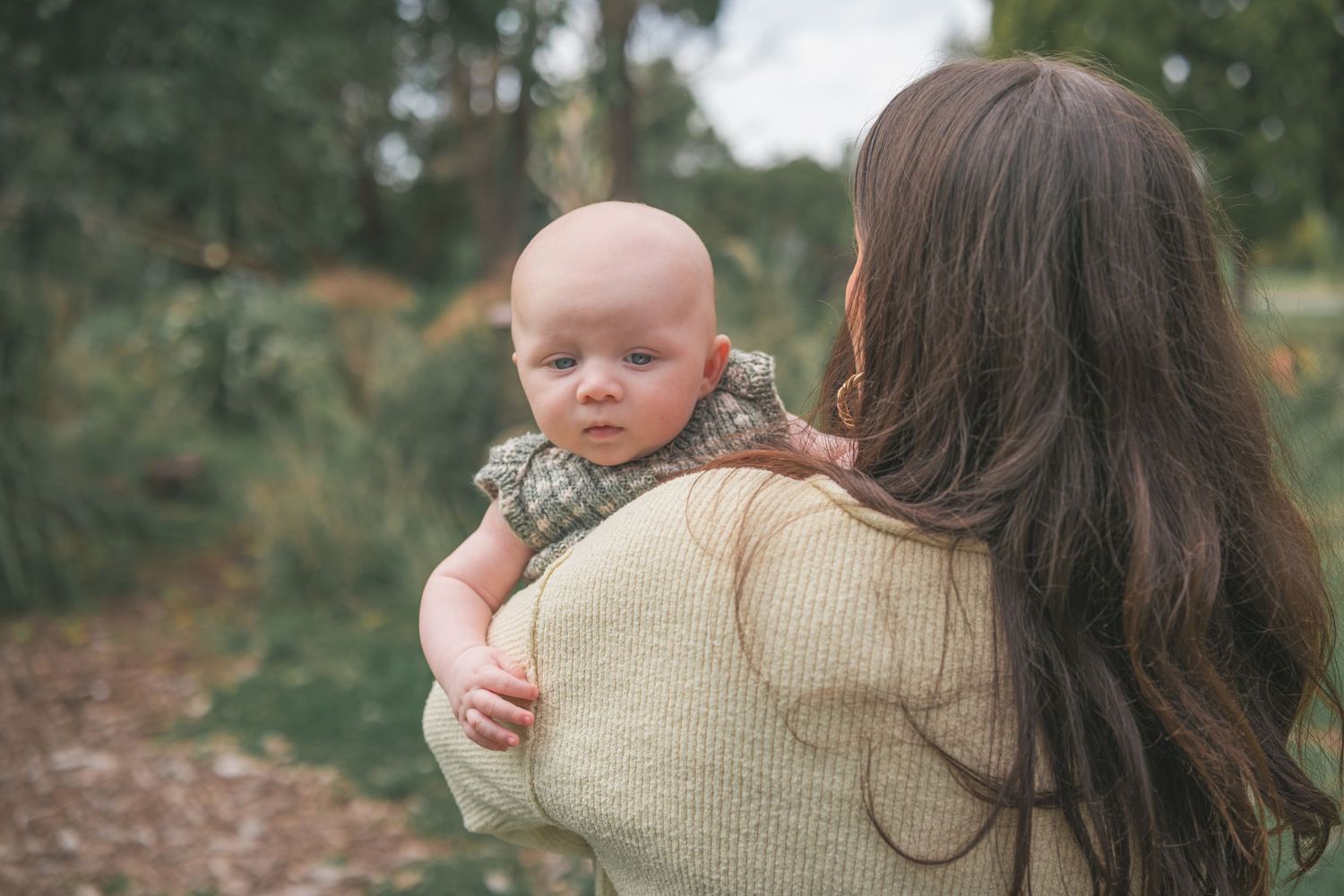 A mother with long brown hair holds her baby over her shoulder outdoors in a lush green park setting.