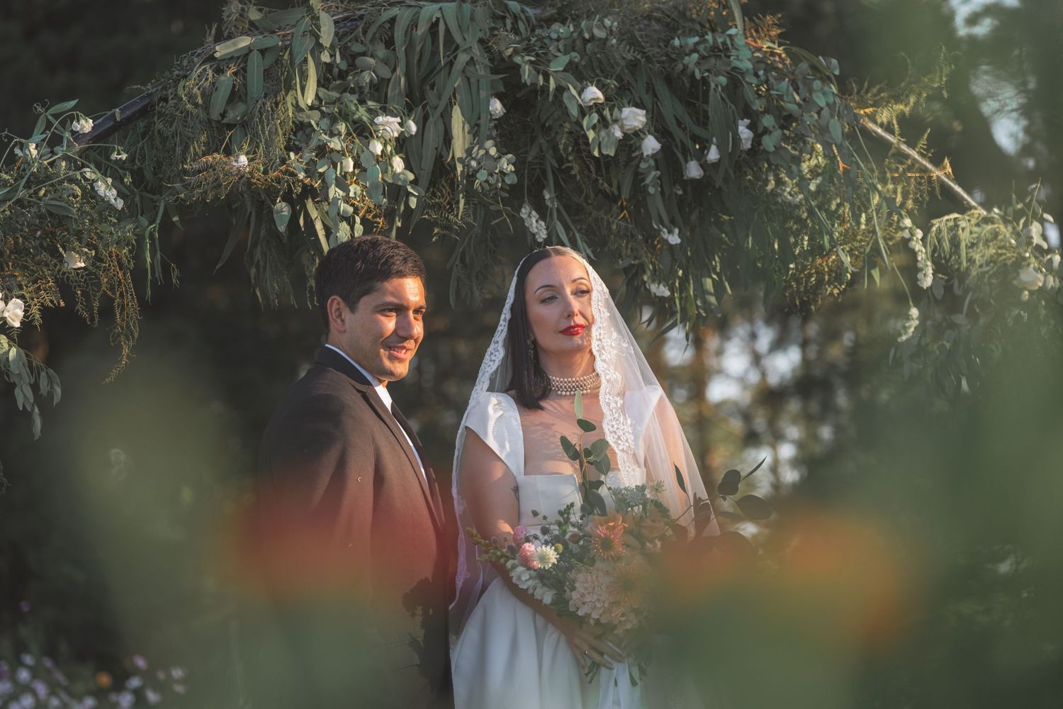 Bride and groom stand under lush greenery arch, bride holding bouquet in soft golden light.