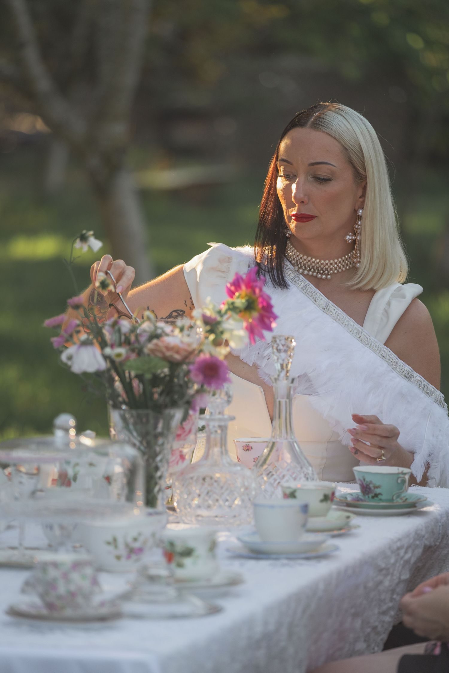 Elegant blonde woman in white dress arranging flowers at an outdoor garden tea party table with fine china.