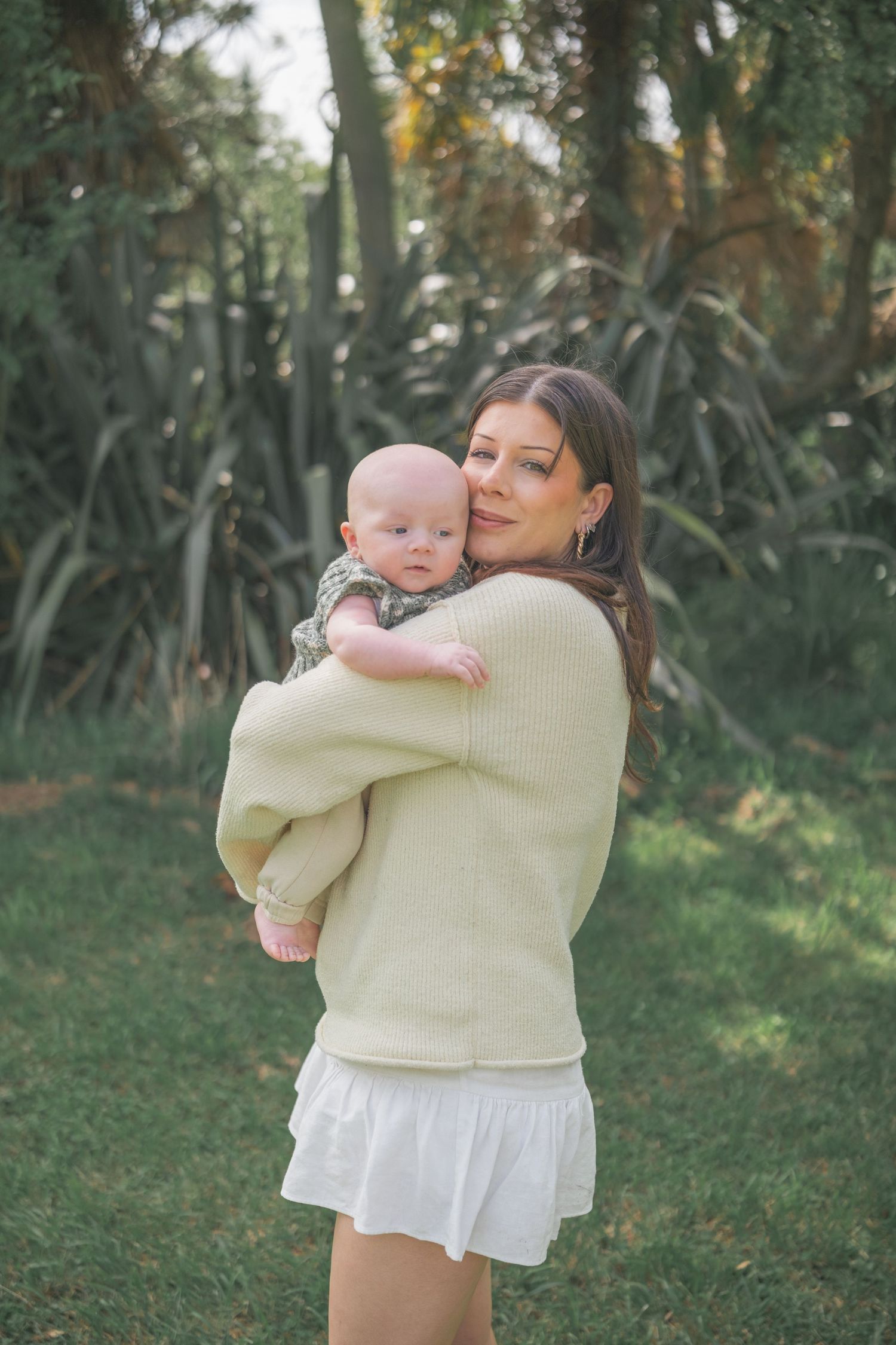 A tender moment between parent and baby in a soft yellow sweater against a green outdoor background.