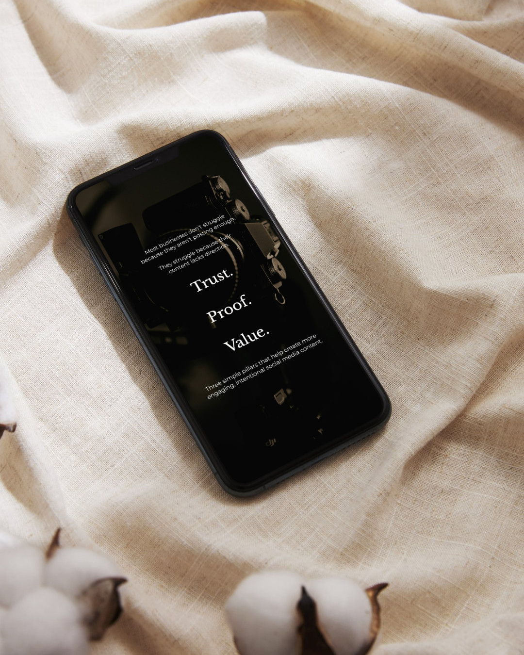 Black smartphone displaying Trust, Proof, Value text on dark screen, resting on cream linen fabric with cotton bolls.