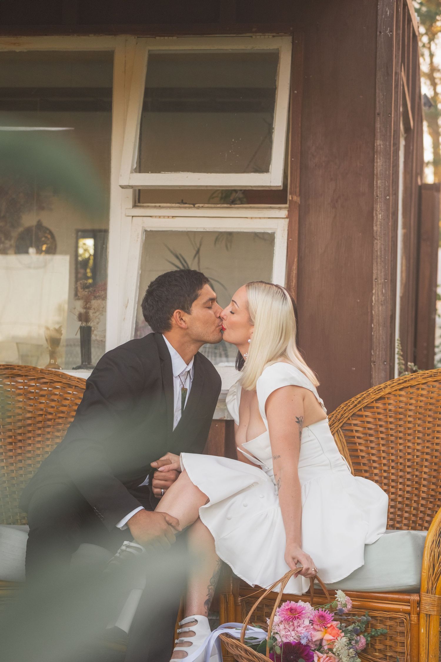 A couple in wedding attire shares a romantic kiss while seated in wicker chairs on a rustic porch.