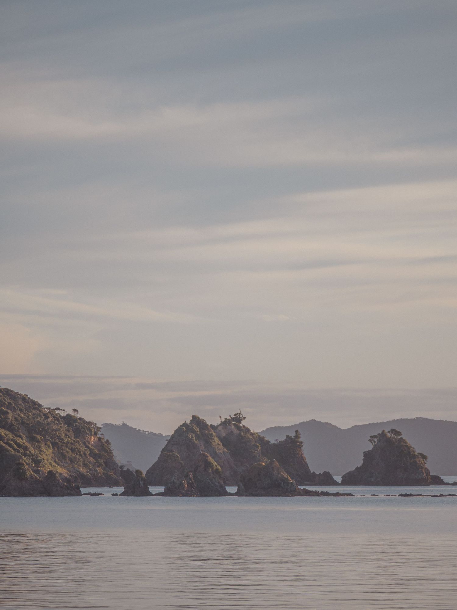 A panoramic coastal landscape showing rocky cliffs and islands emerging from calm waters under an overcast sky.