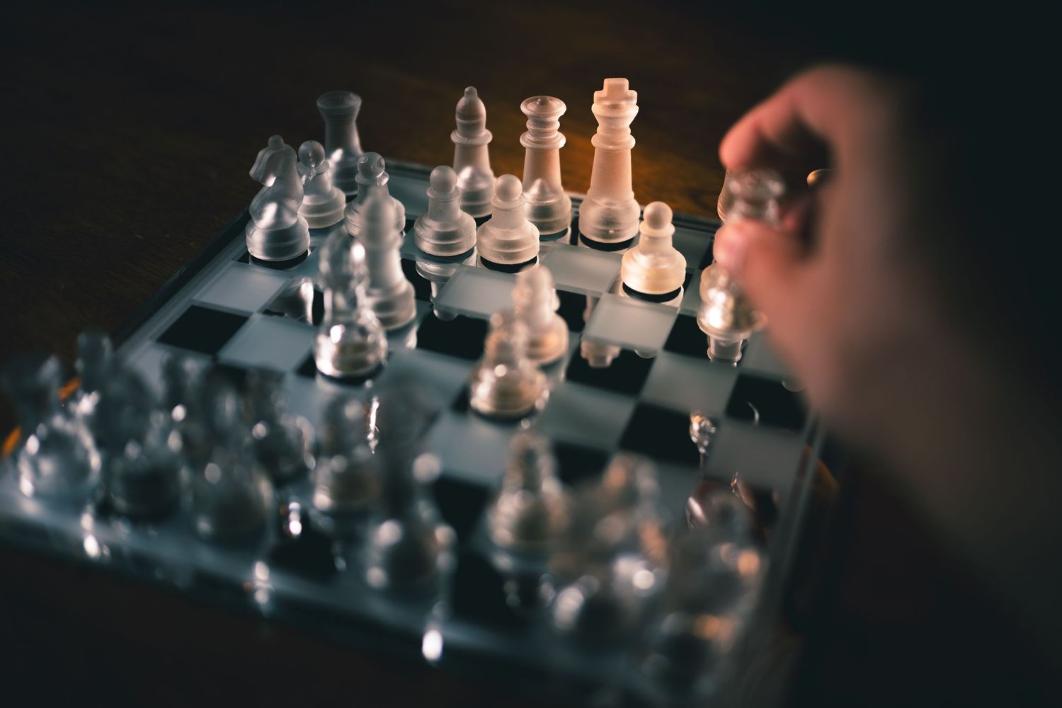 A hand moves a chess piece on a glass chess board in a dramatic, dark-lit strategic game setting.