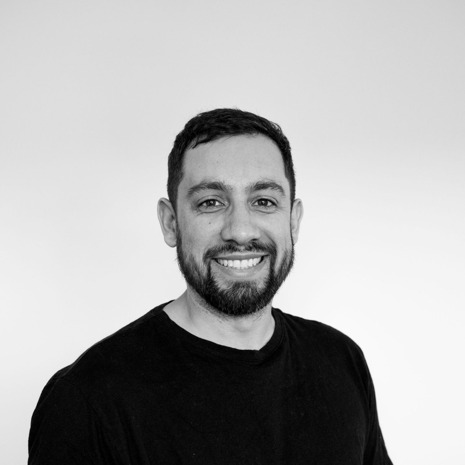 Smiling man with beard wearing black t-shirt poses for professional headshot photo against white background.