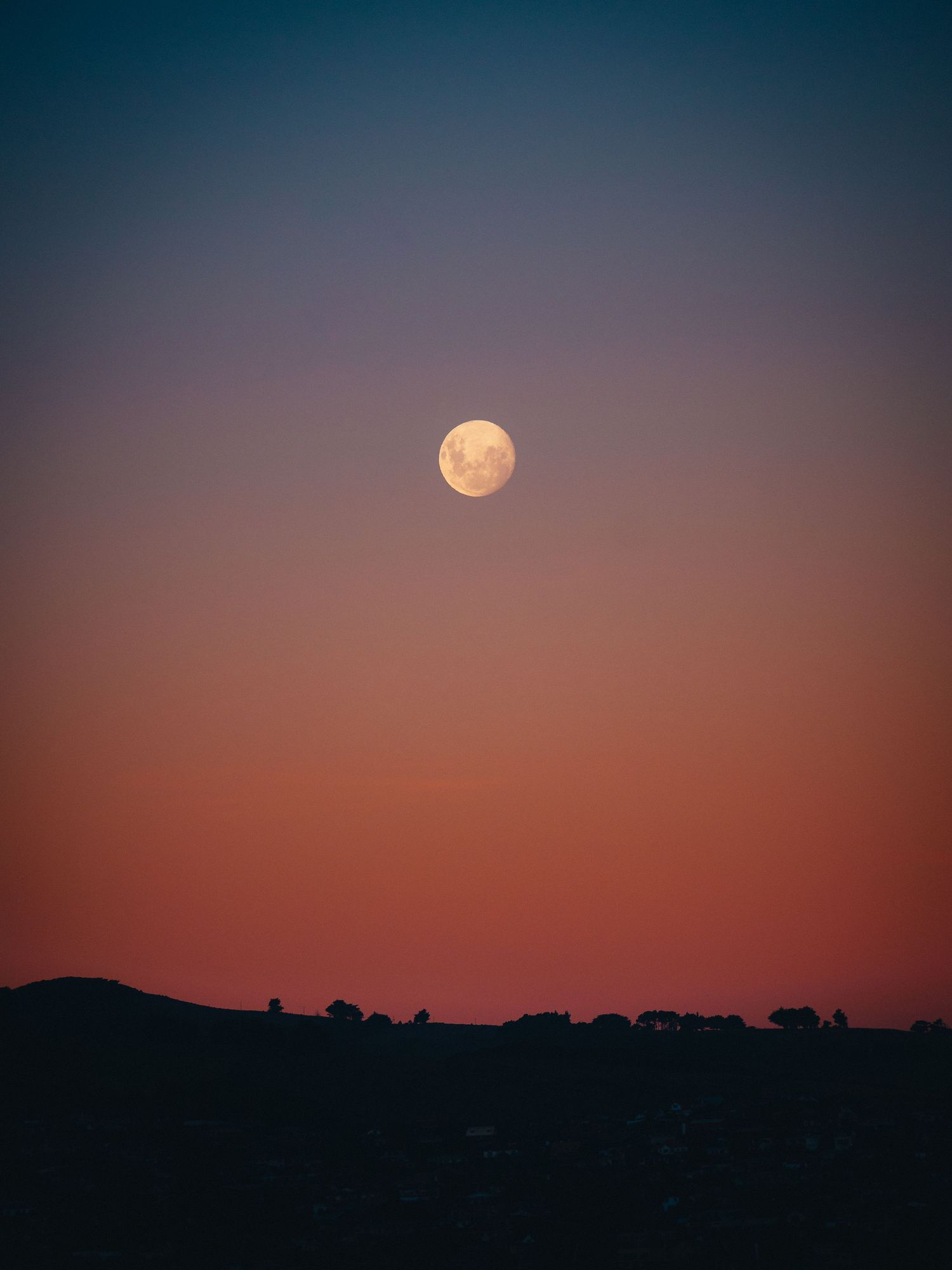 Full moon glowing in a gradient twilight sky over dark silhouetted hills with trees on the horizon.