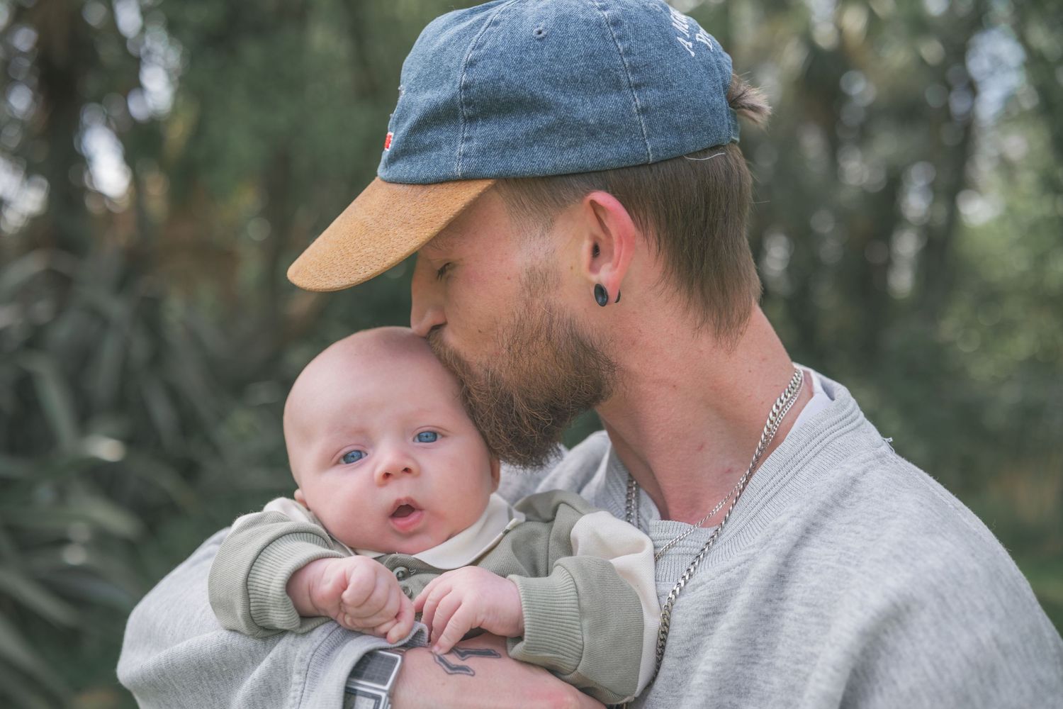 A parent wearing a baseball cap holds and kisses a small baby in a series of tender outdoor moments.