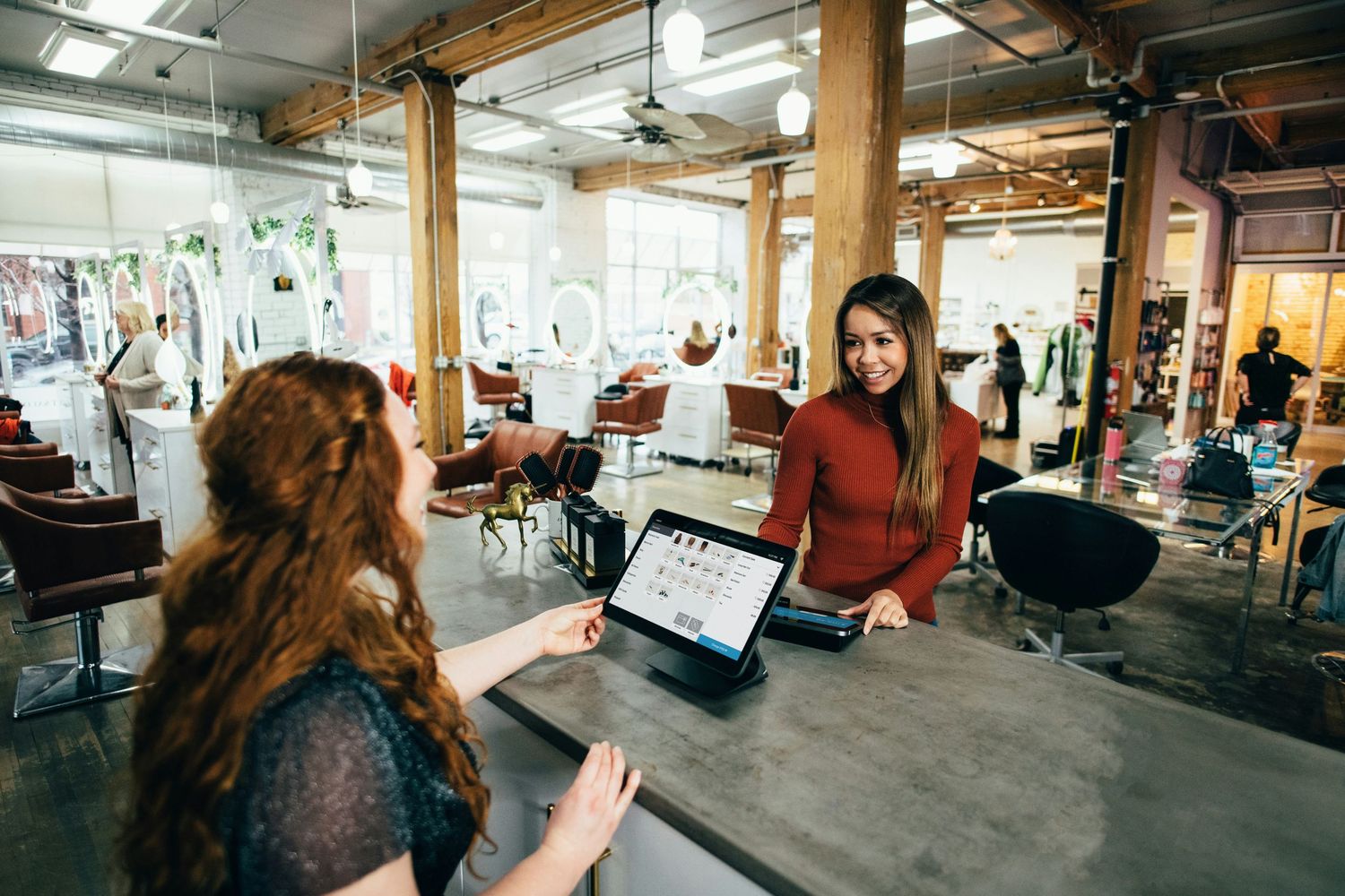 Two women interact at a salon reception desk with a tablet POS system in a modern hair salon.