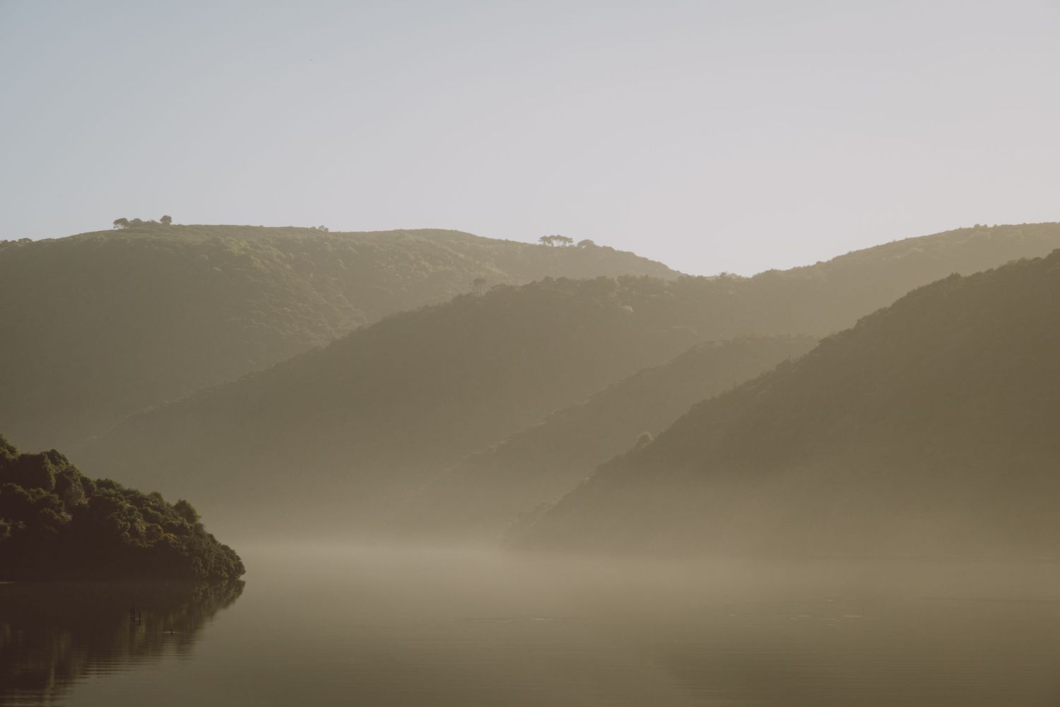 Misty morning landscape with rolling hills silhouetted against a hazy sky reflected in calm waters below.