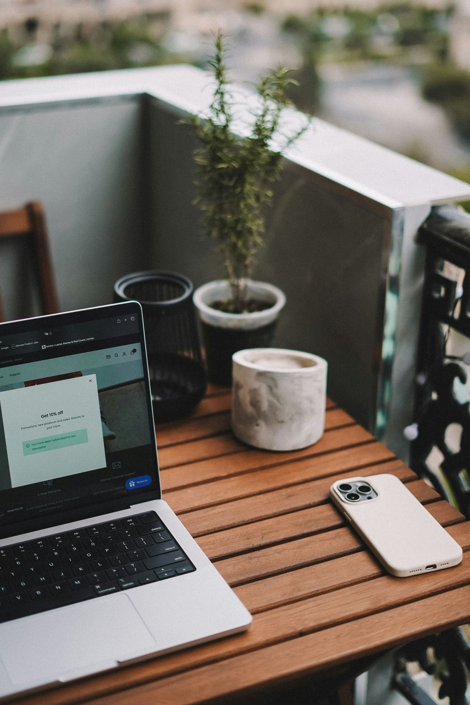 Laptop and smartphone on wooden balcony table with coffee cup and potted plant in urban outdoor workspace.